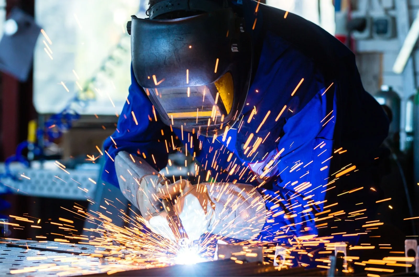 A person wearing a welding helmet and blue protective clothing welding metal, with sparks flying around in a workshop.