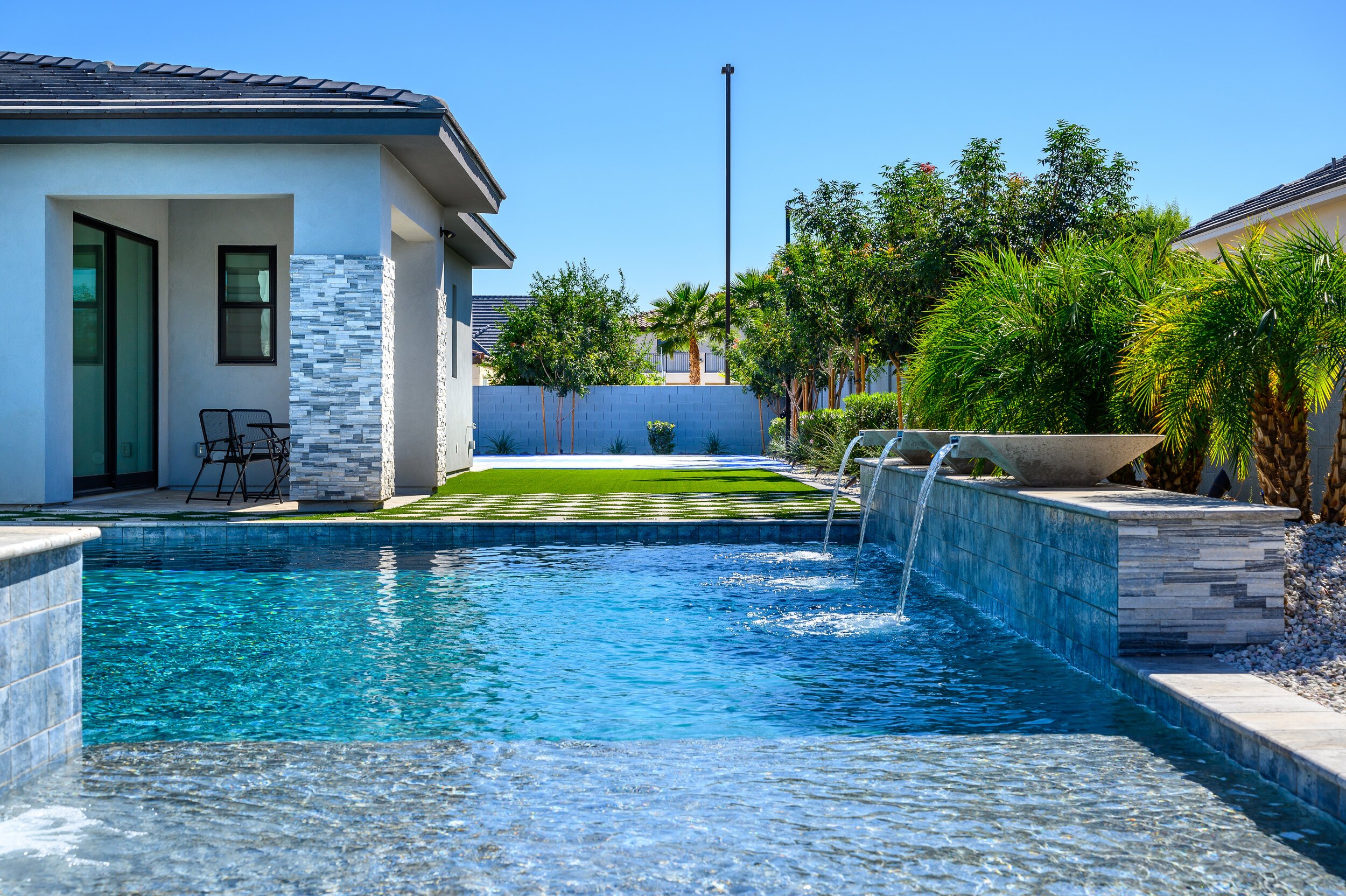 Modern backyard with a swimming pool, grassy area, and tropical plants under a clear blue sky.