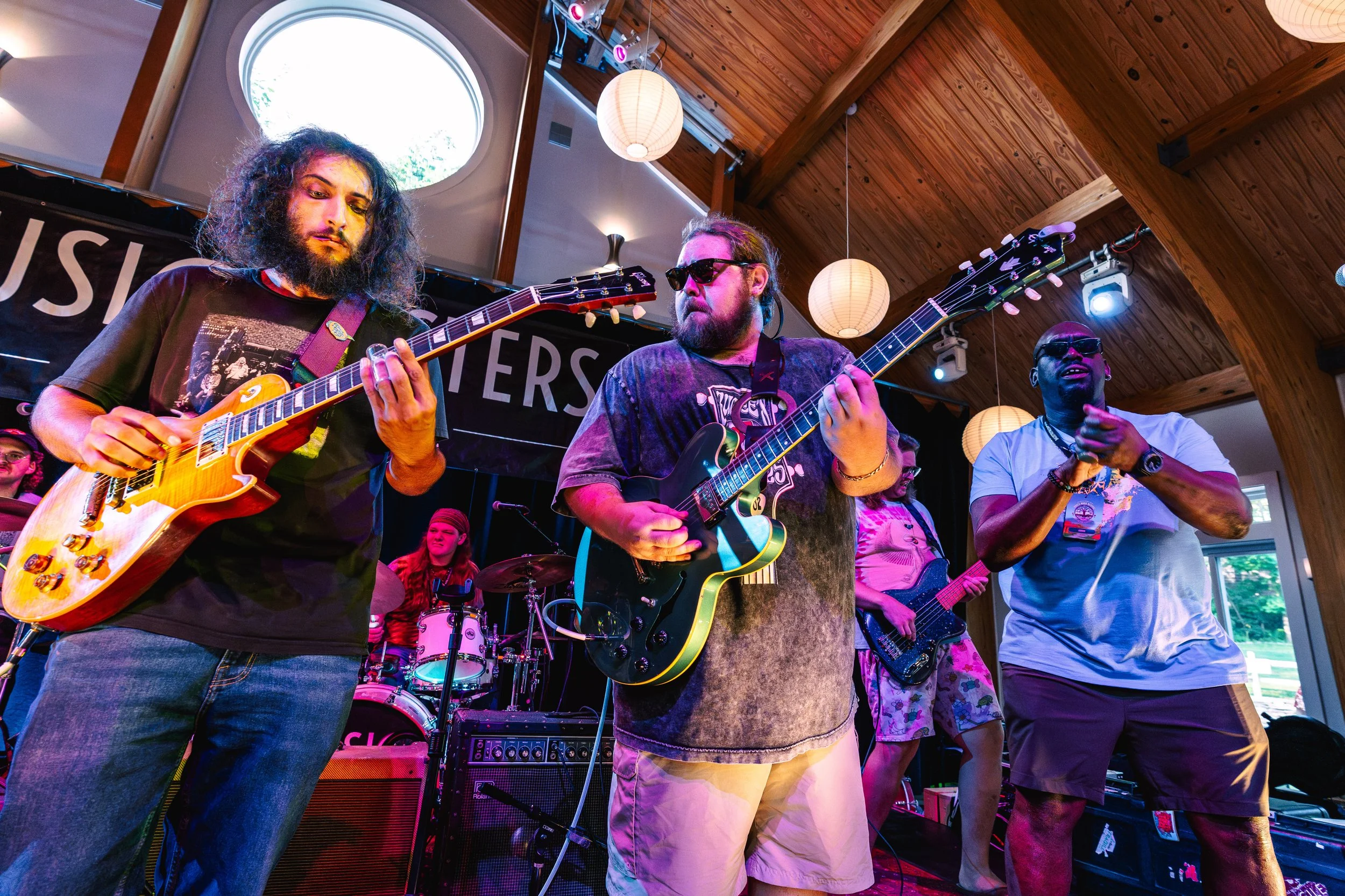 A band of five musicians performing on stage at an indoor venue with wooden ceiling and paper lantern lights. They include two guitar players, one on the left with curly hair and a beard, one in the center with sunglasses, and a bass player on the right. In the background, a female drummer and a female bass player are visible. The stage has a black banner with partially visible text.