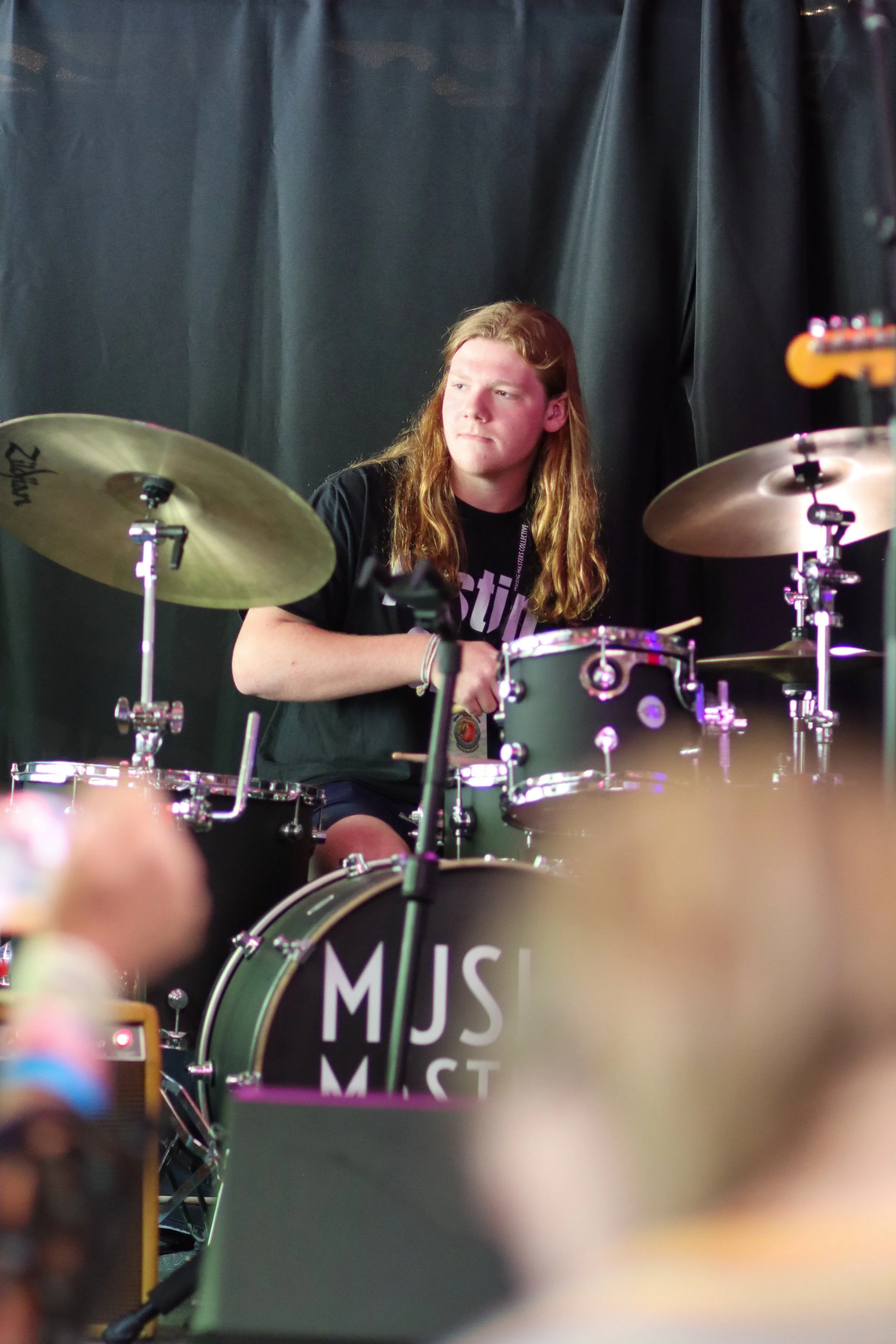 A young person with long red hair playing a drum set during a performance, with a black curtain in the background.