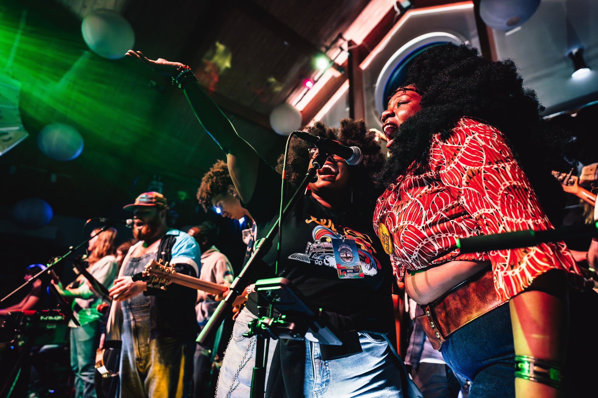 Band performing live on stage with three musicians, one playing piano, another playing banjo, and the third playing acoustic guitar. The stage is decorated with paper lanterns and blue lighting.