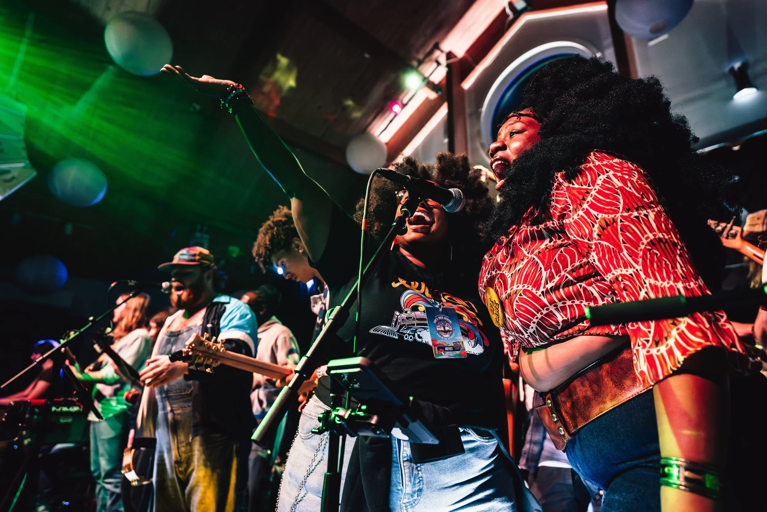 A group of musicians and singers performing on stage, with two women in the foreground singing into microphones. One woman is wearing a red patterned shirt and the other a black T-shirt with a colorful graphic. Other performers are visible in the bac