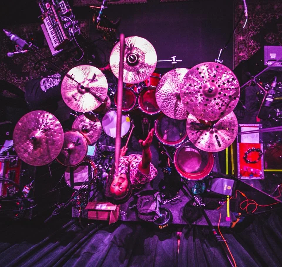 Overhead view of a female drummer playing a drum set on stage with various equipment, illuminated by pink and purple stage lights.