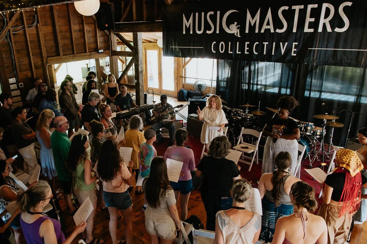 A group of people gathered in a wooden music venue, listening to a woman speaking in front of a stage with musical instruments, under a large banner that reads 'Music Masters Collective'.