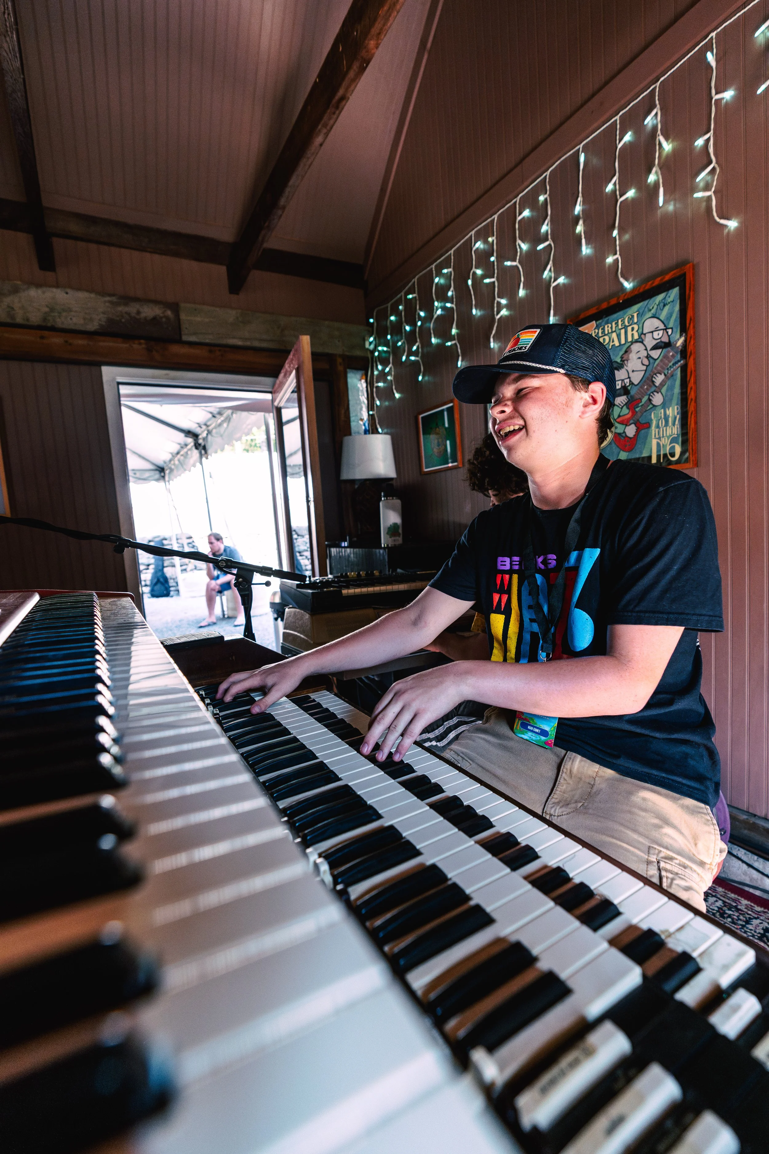 Young person playing a keyboard and smiling indoors with string lights on the wall and a poster behind them, with an open door showing outdoor seating.