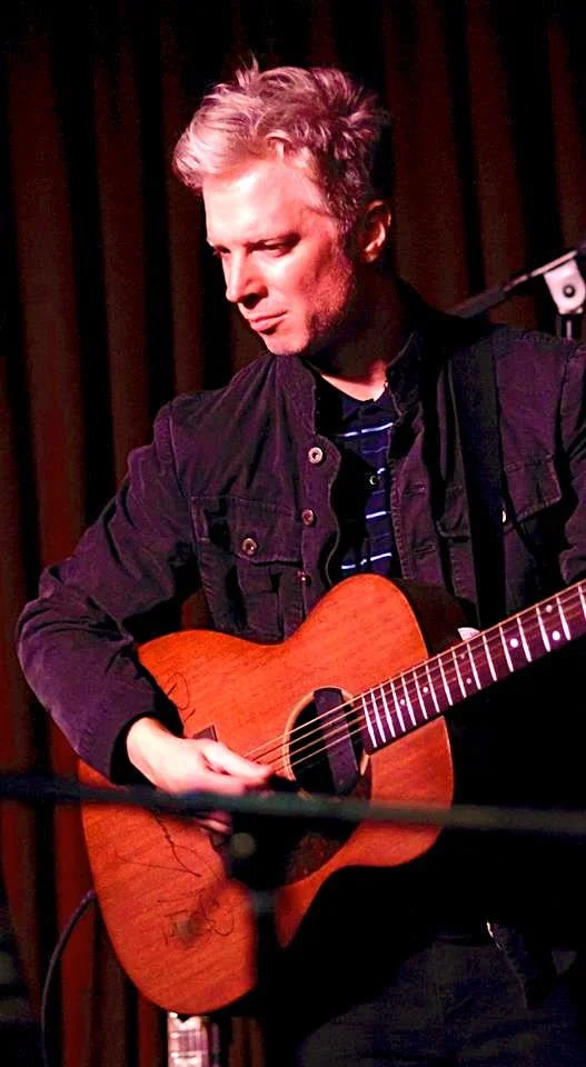 A man playing an acoustic guitar on a stage with a dark curtain background.
