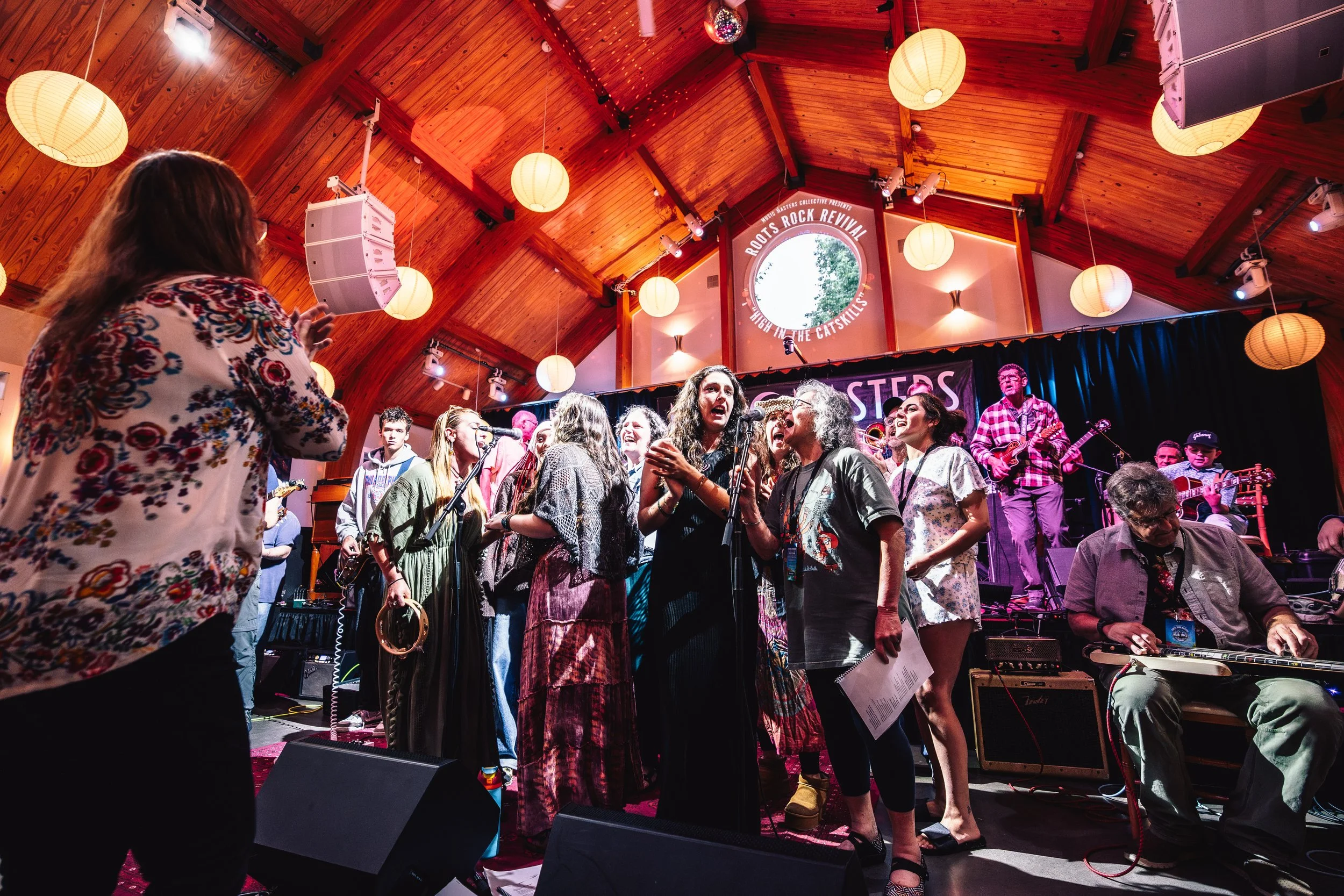 A group of musicians and singers performing on stage in a wooden-ceiling concert hall with paper lantern lights hanging from above. There are people singing into microphones, playing instruments, and enjoying a lively musical event.
