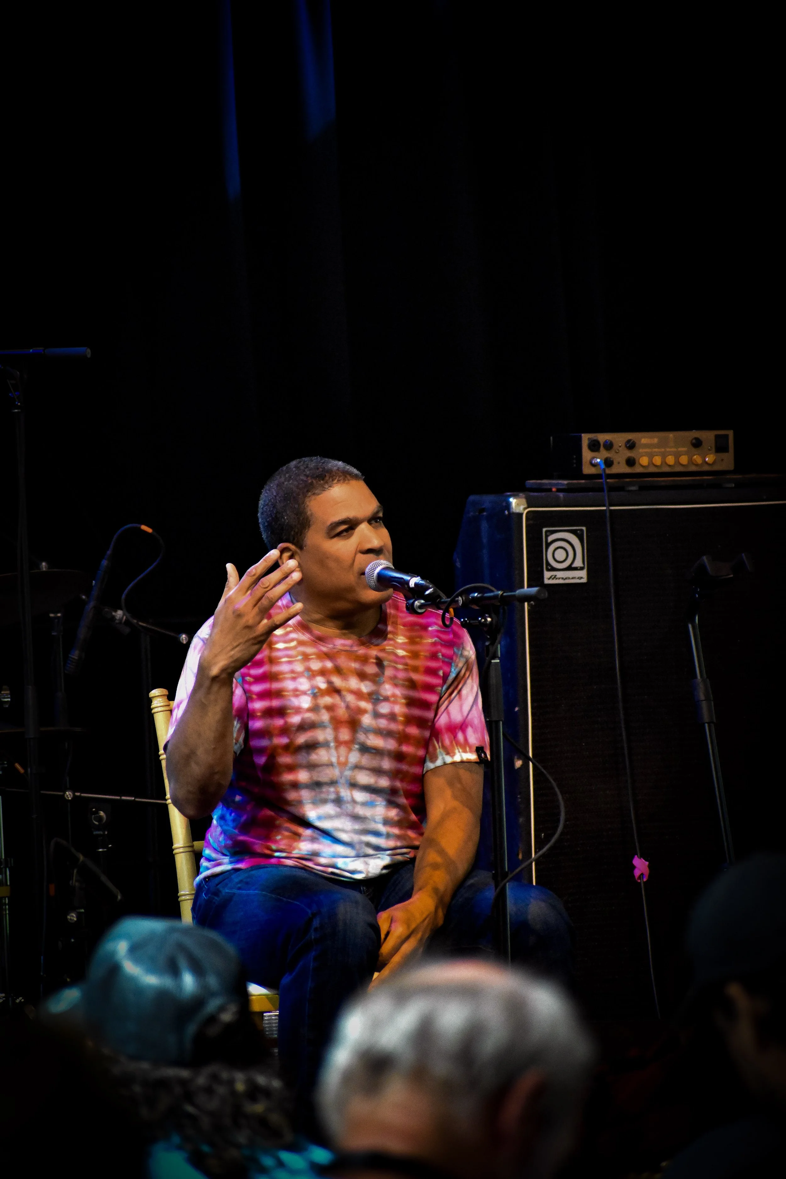 Musicians performing on stage with guitars and drums, colorful lighting, and a black curtain backdrop.
