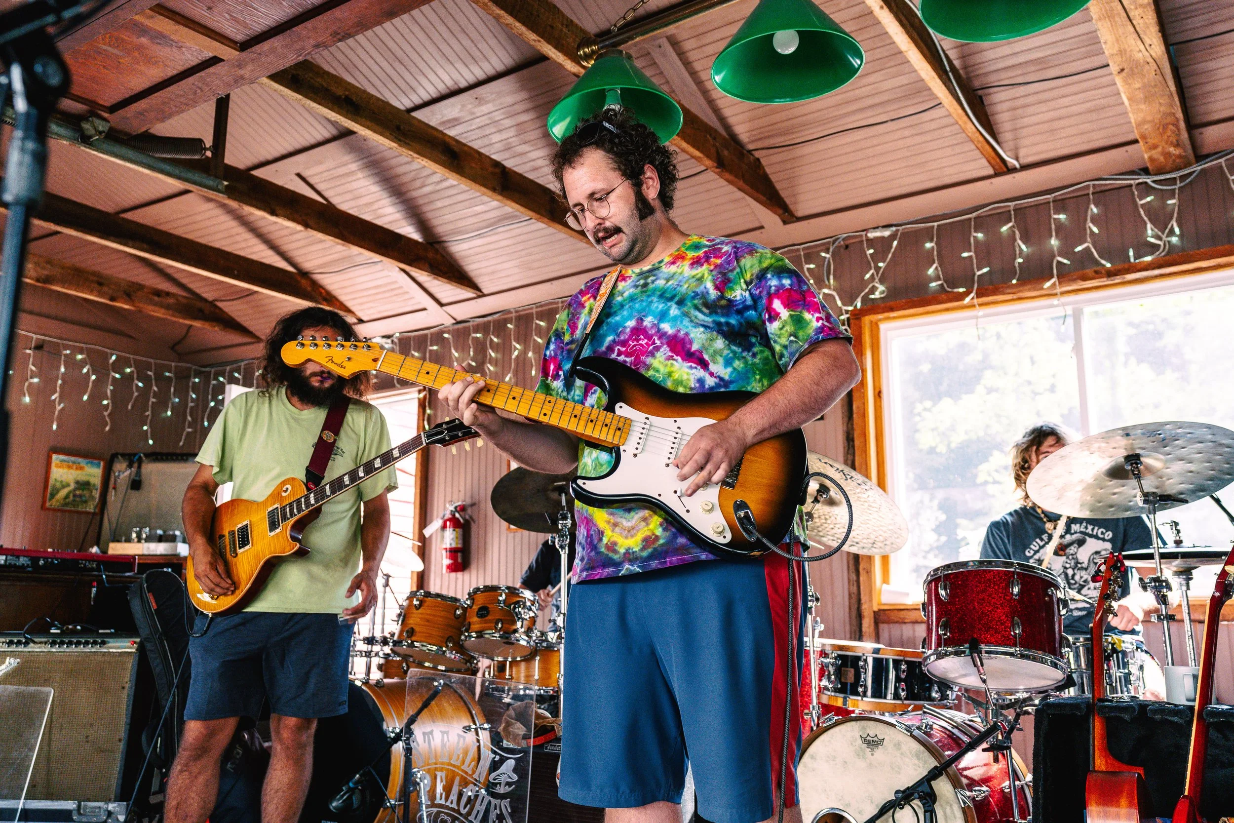 A band of four musicians playing instruments in a rustic indoor setting with string lights and large windows.