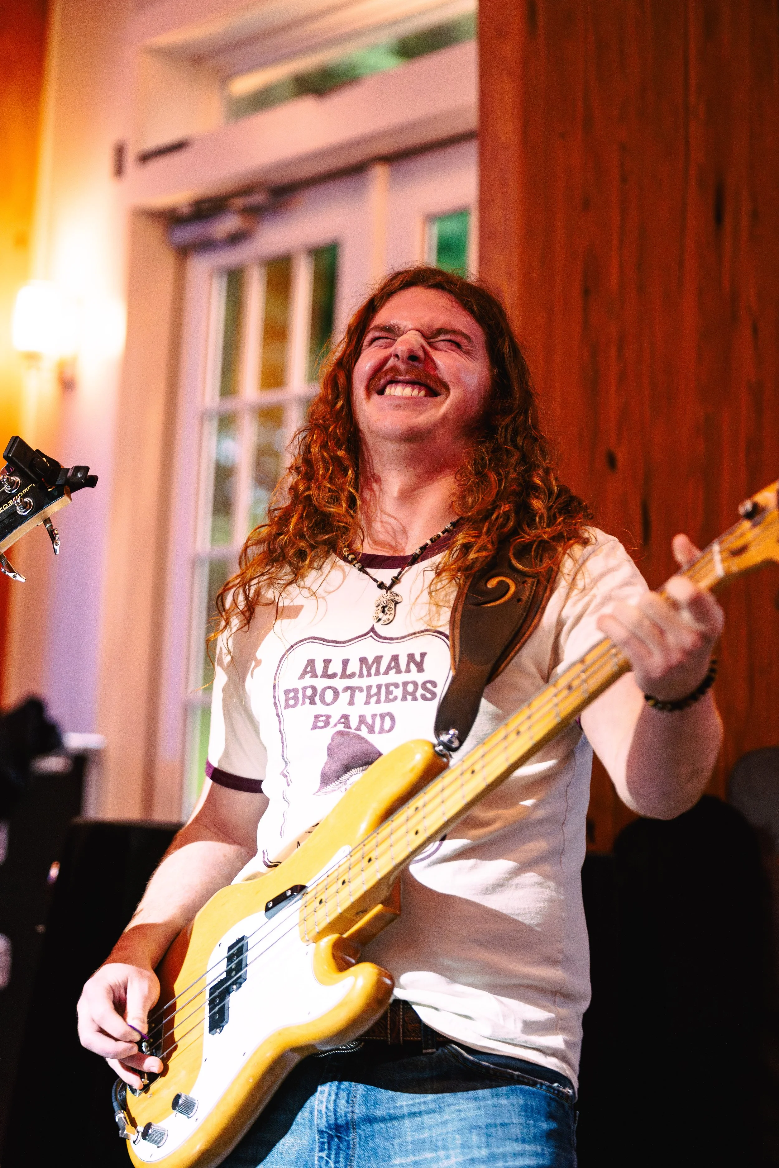 A man with long curly hair smiling while playing a bass guitar indoors.