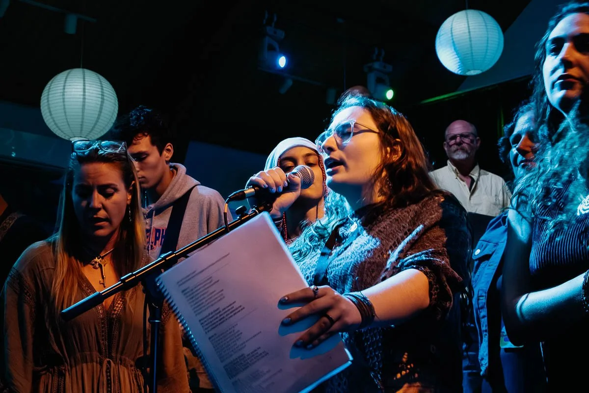 A woman with glasses and red hair speaks into a microphone, holding a printed sheet, surrounded by several people on a stage with hanging paper lanterns and stage lights.