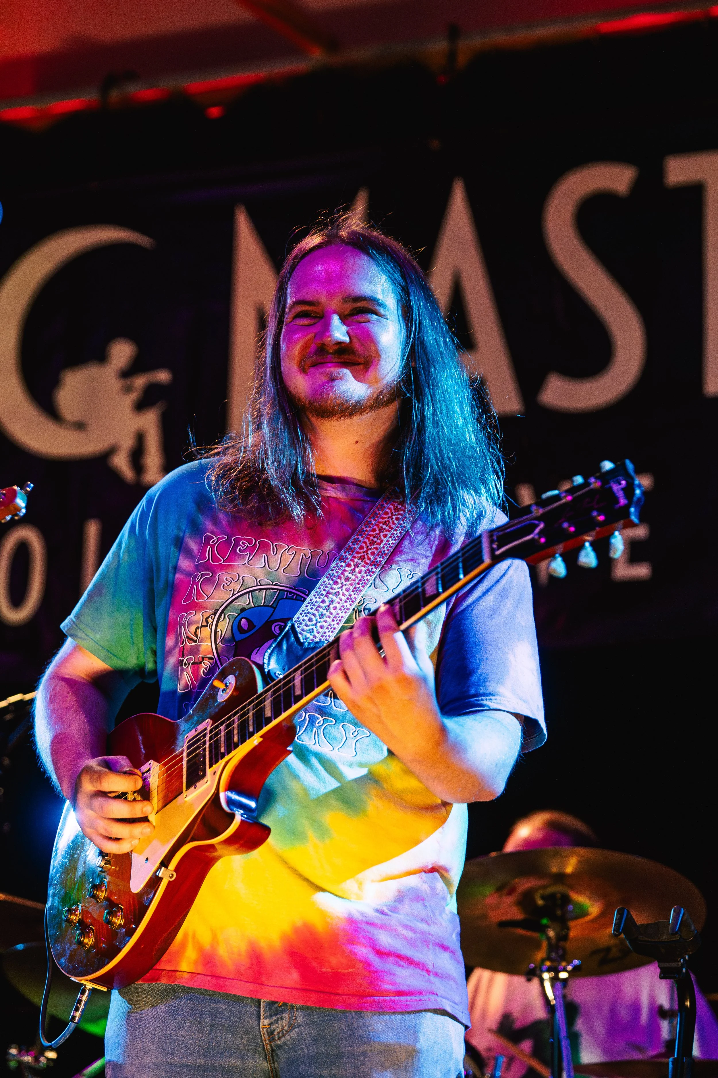 A man with long hair playing an electric guitar on stage, wearing a colorful tie-dye T-shirt, with a drum set in the background.