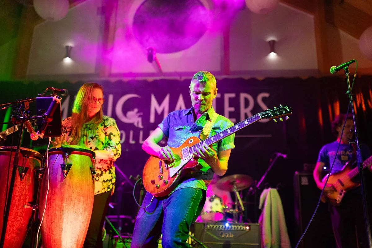 Musicians performing on stage with colored lighting, including a guitarist, a woman playing conga drums, and a bass player, with a backdrop reading 'Music Matters'.