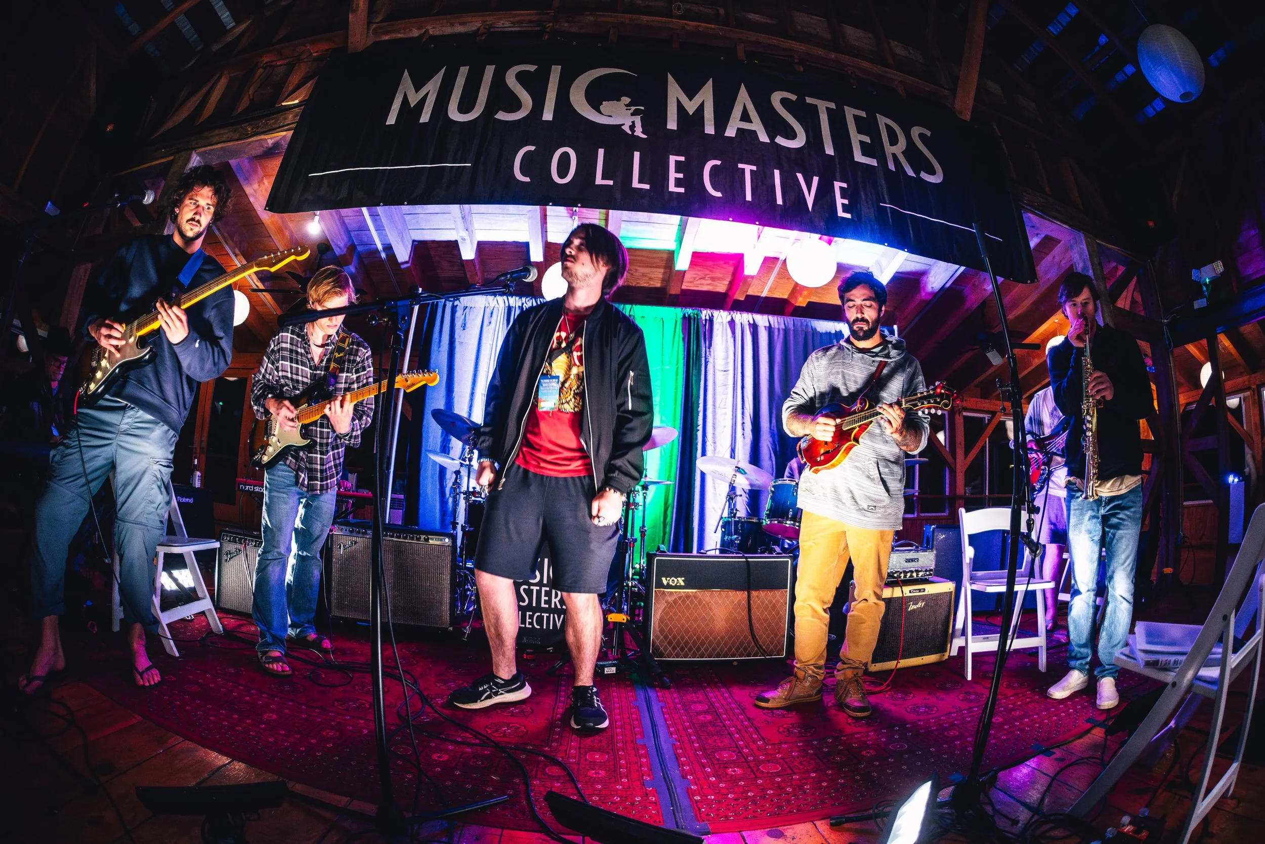 A band performing live on stage at Music Masters Collective event, with six musicians playing various instruments including electric guitars, bass guitar, drums, and saxophone. The stage features colorful lighting and a banner overhead that reads "Mu