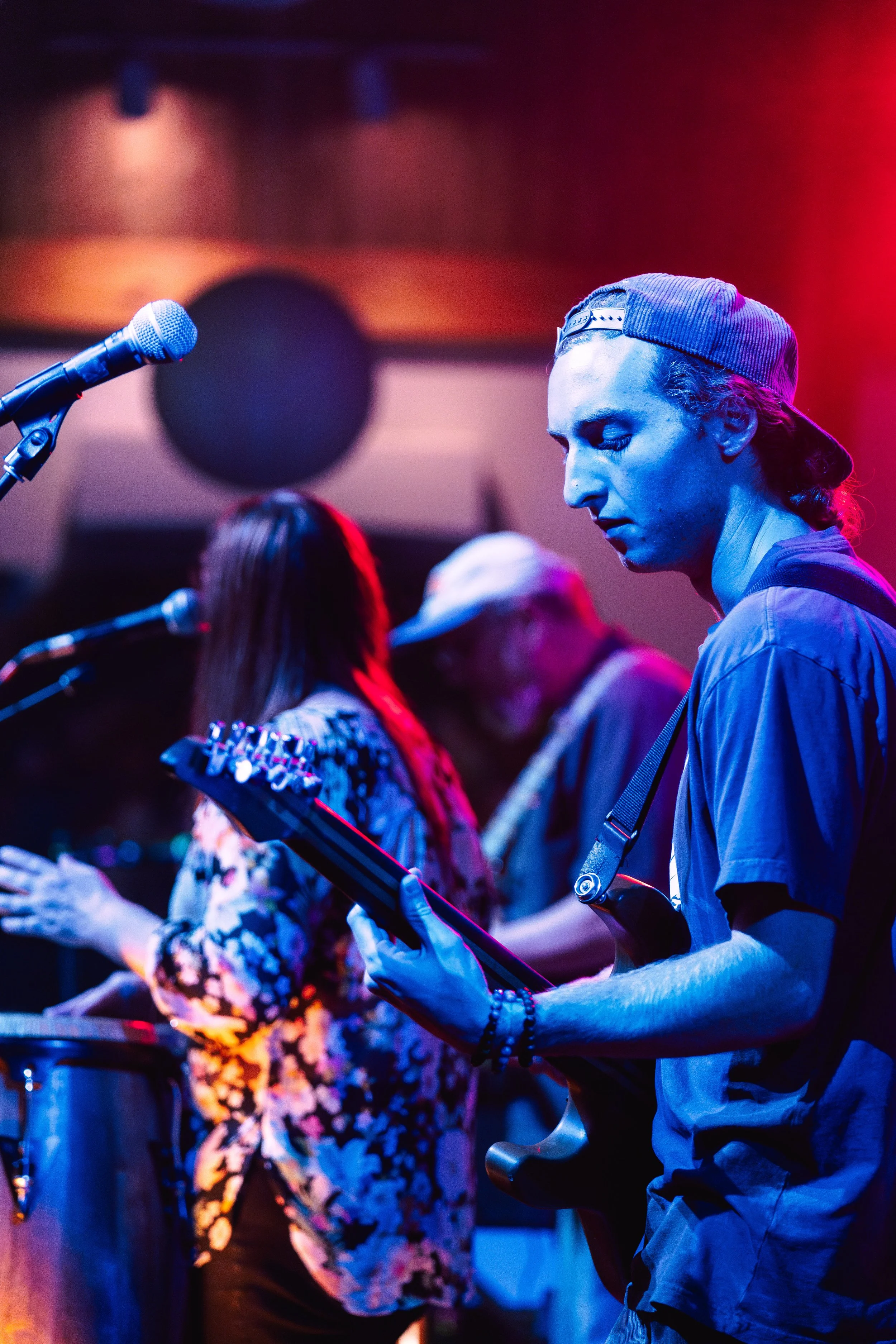 Three musicians playing instruments on stage under colorful lights, including a young man with a backwards cap playing an electric guitar.