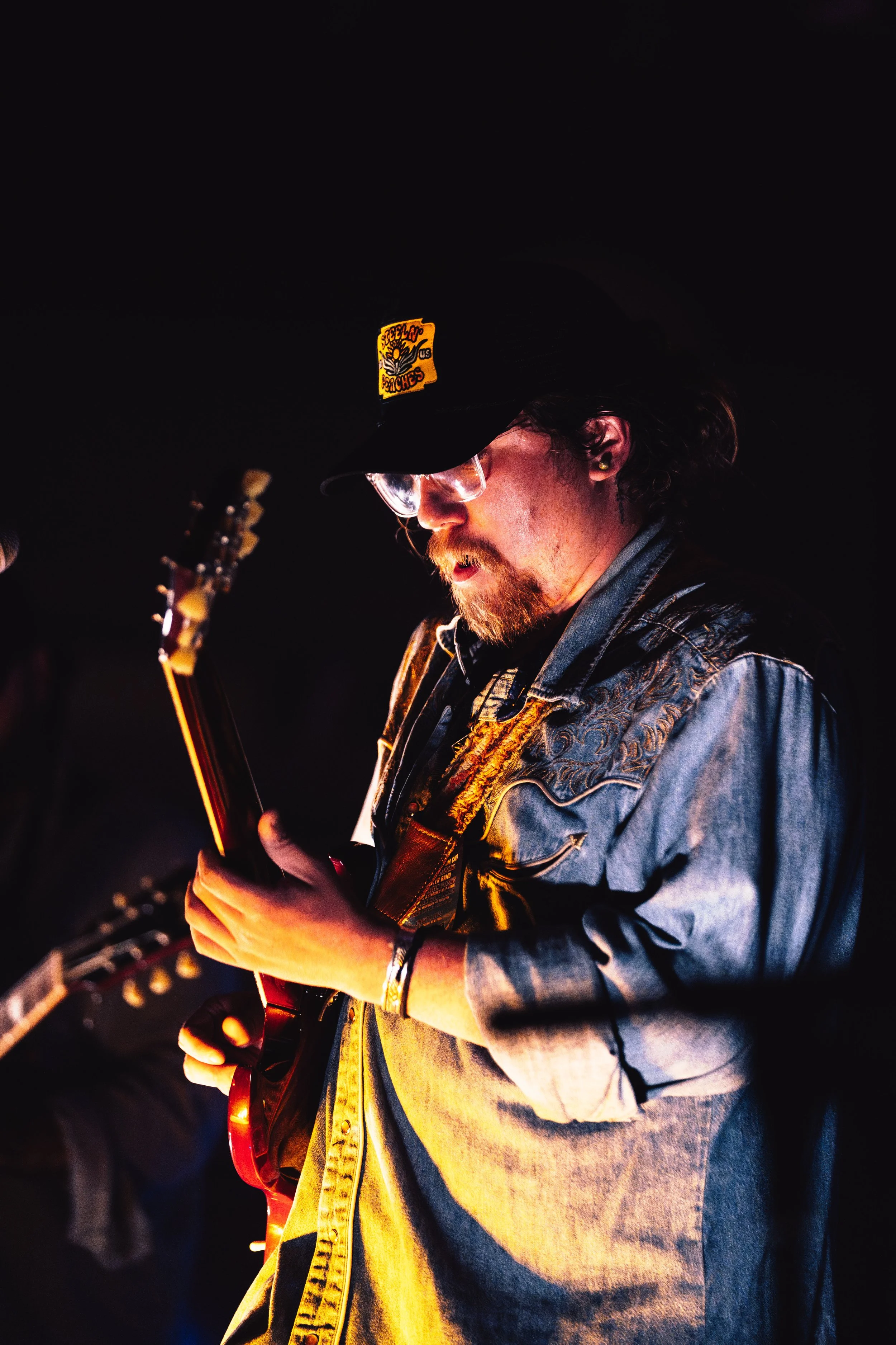 A man in a denim shirt, wearing a black cap and glasses, playing an electric guitar in a dark setting with warm lighting.