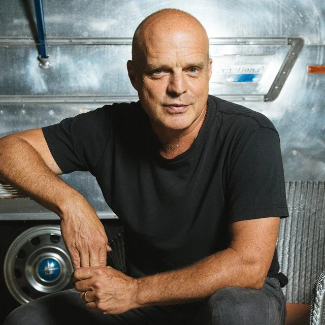 A bald man in a black t-shirt sitting in front of a metallic background, resting his arm on his knee and looking at the camera.