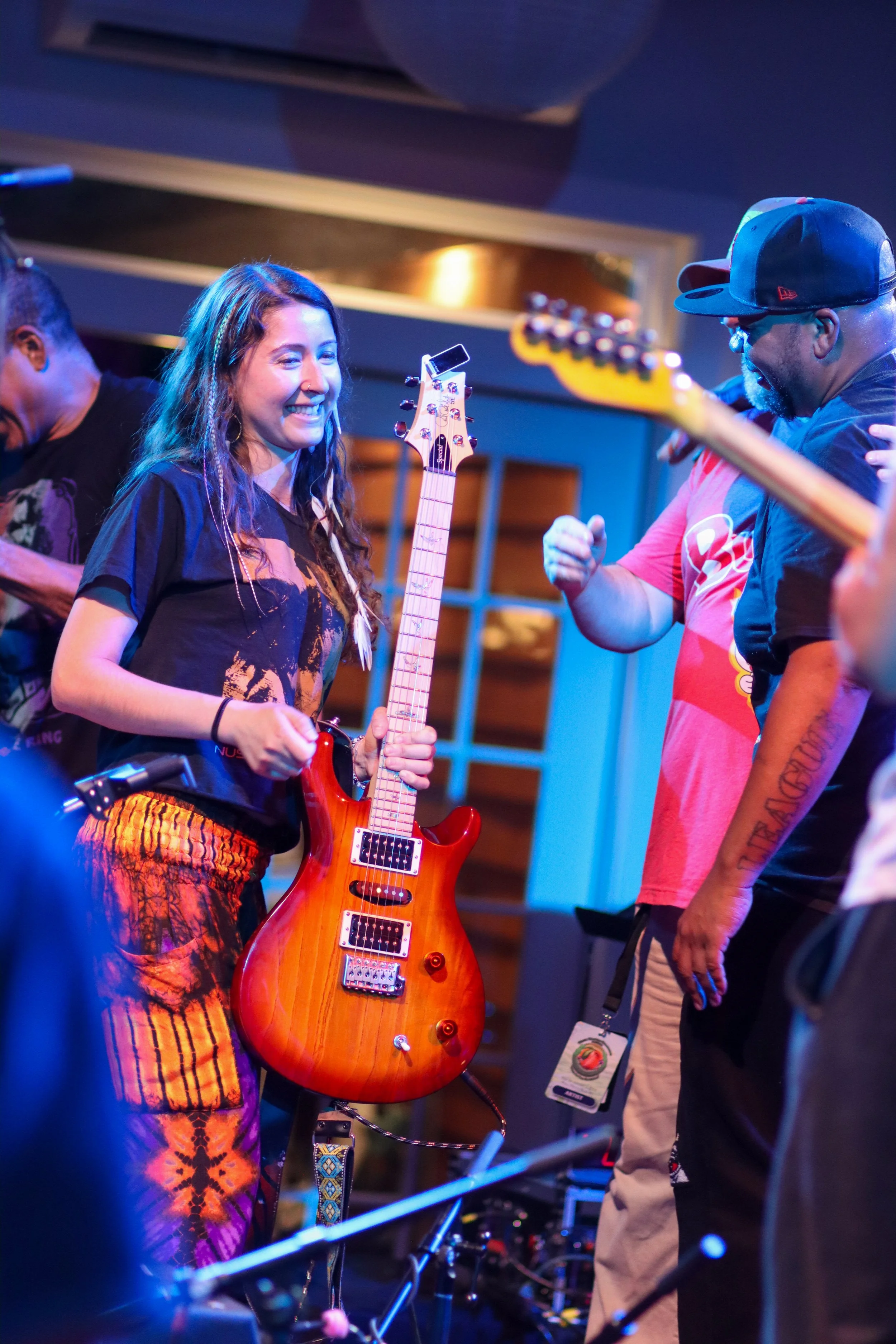 A young woman smiling and holding an electric guitar in a performance setting, surrounded by other musicians.