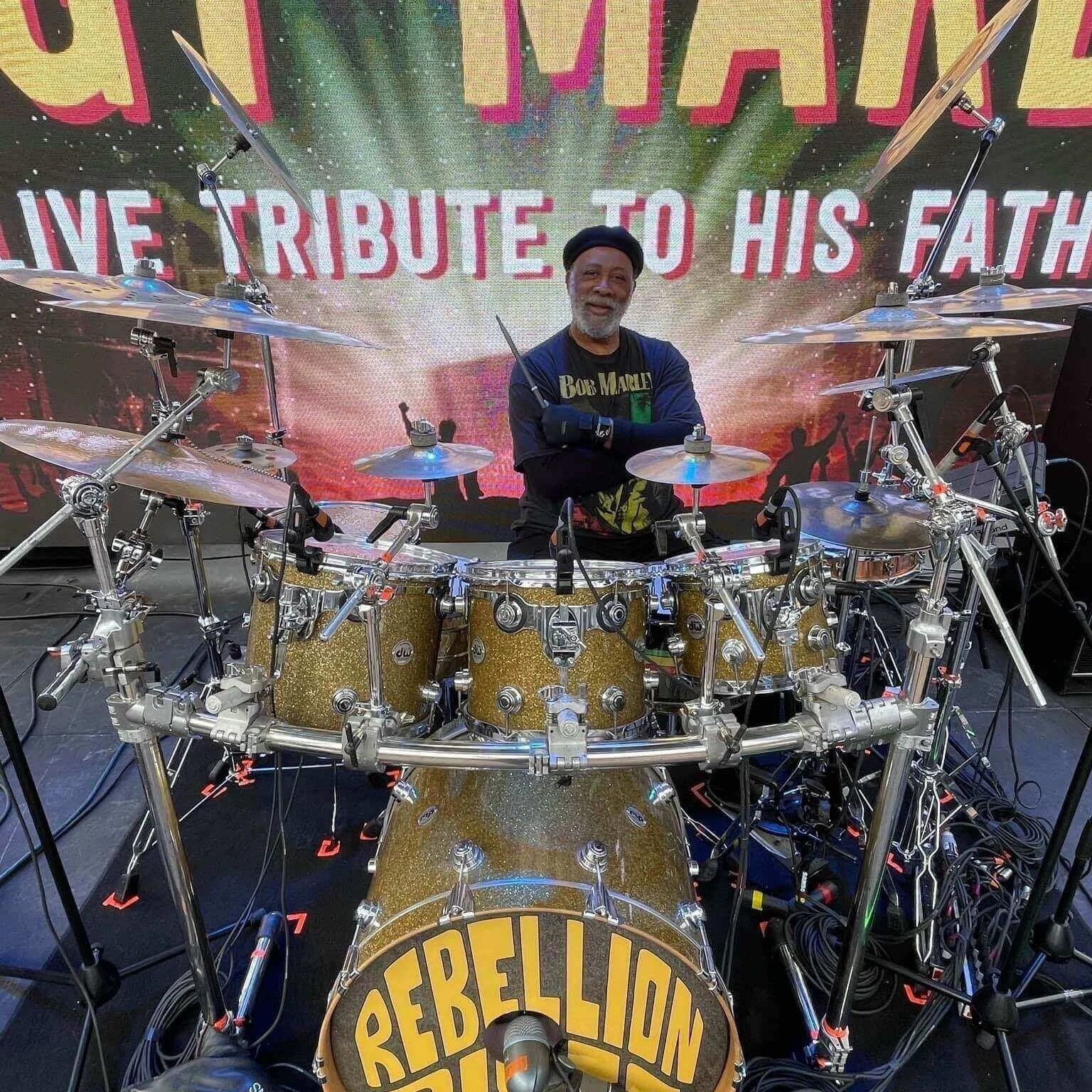 A man sitting behind a gold drum set with the word 'REBELLI0N' painted on the bass drum. He is smiling and wearing a Bob Marley t-shirt and a black cap, with a large digital screen behind him displaying a message to his father, reading 'LIVE TRIBUTE TO HIS FATHER.'