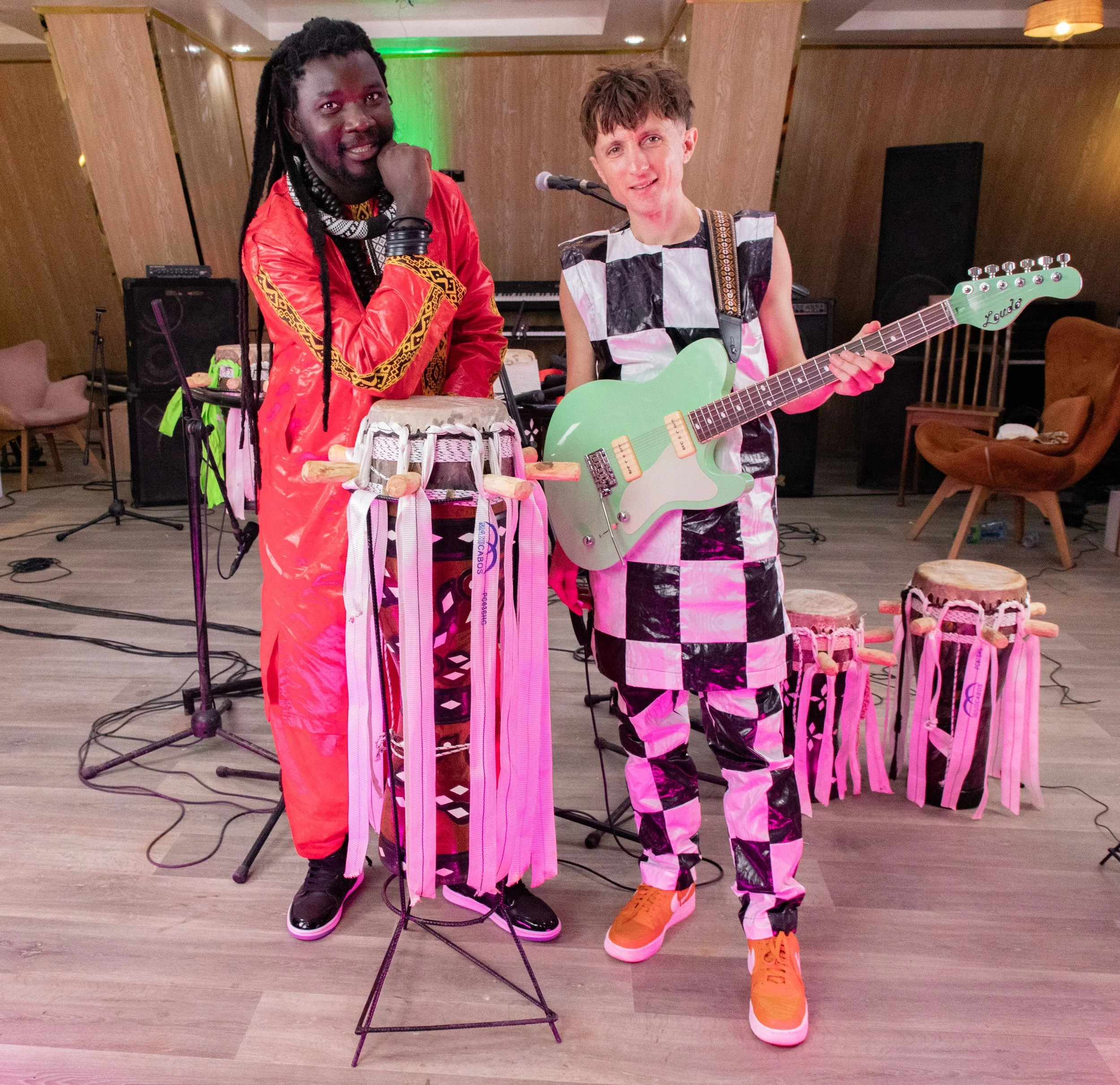 Two musicians standing together in a performance space, both with musical instruments. One is wearing a red outfit and holding a drum, the other is wearing a black and white checkered outfit and holding a pale green electric guitar.