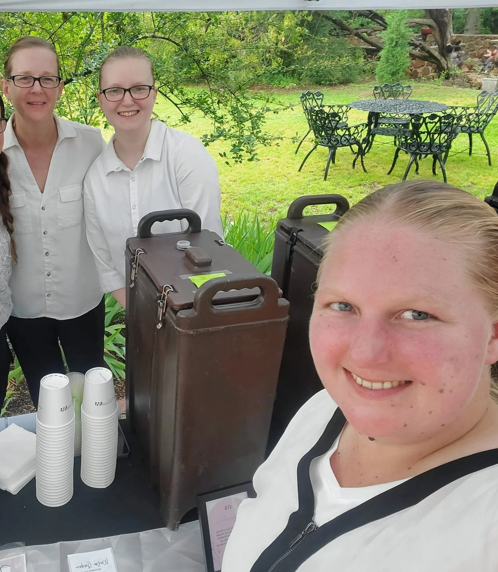 A group of smiling women at an outdoor event, with a woman in the foreground taking a selfie. They are standing behind a table with brown beverage dispensers, cups, and napkins, in a garden setting with trees and a patio table in the background.