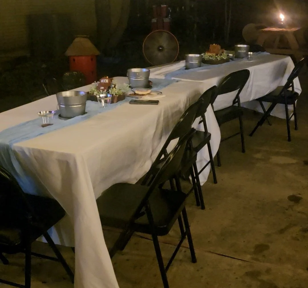 A long rectangular table set for a gathering, covered with a white tablecloth and decorated with small flower arrangements, metal buckets, and rustic decorations in an outdoor or semi-outdoor setting during nighttime.
