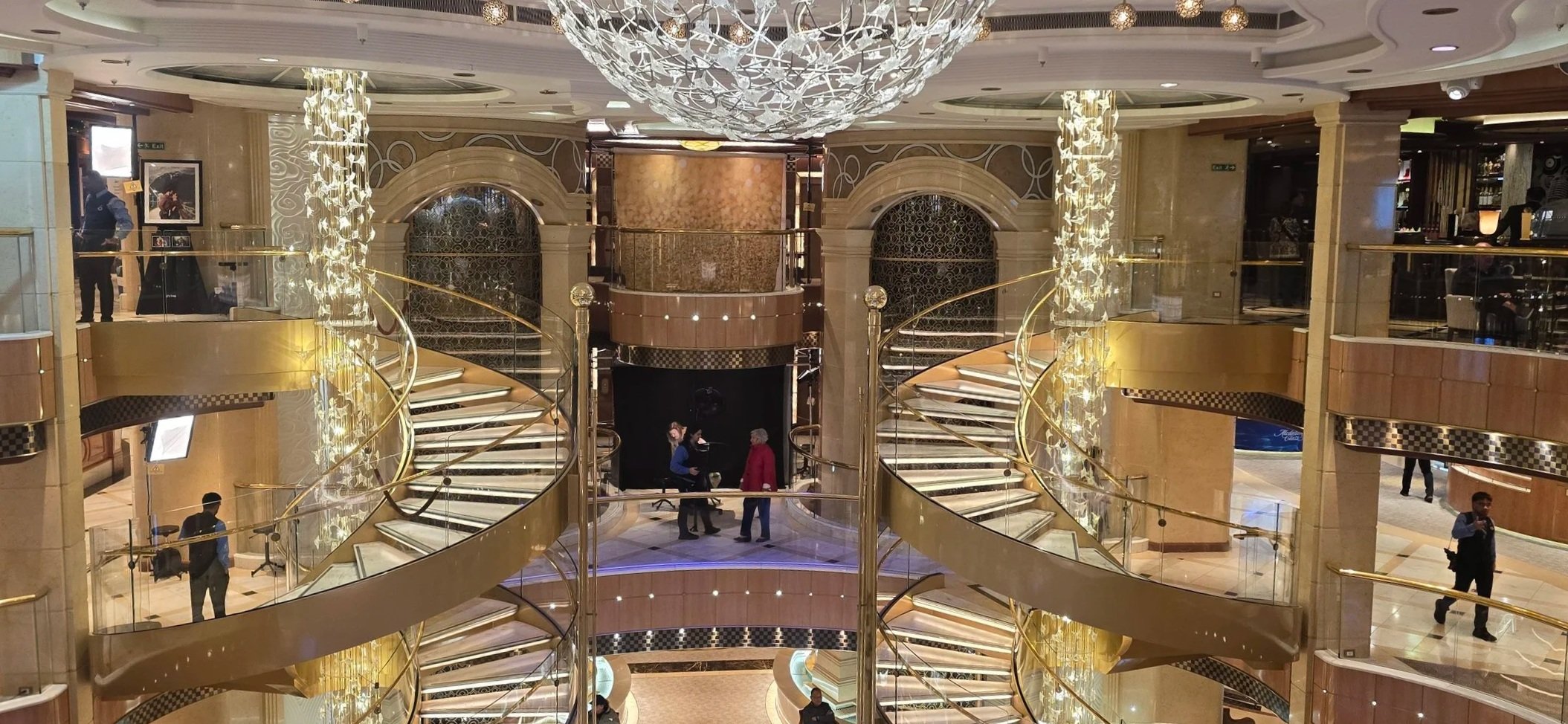 Interior of a luxurious hotel lobby with grand staircase, ornate chandeliers, and gold accents.