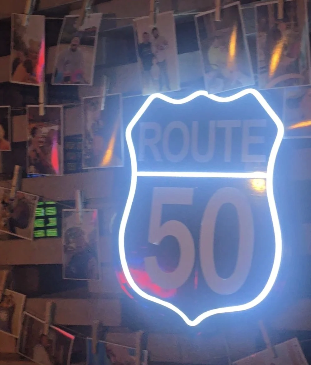 Neon sign shaped like a US highway shield with the text 'ROUTE 50'. Behind the sign, there are photos clipped onto strings with clothespins, some showing people.