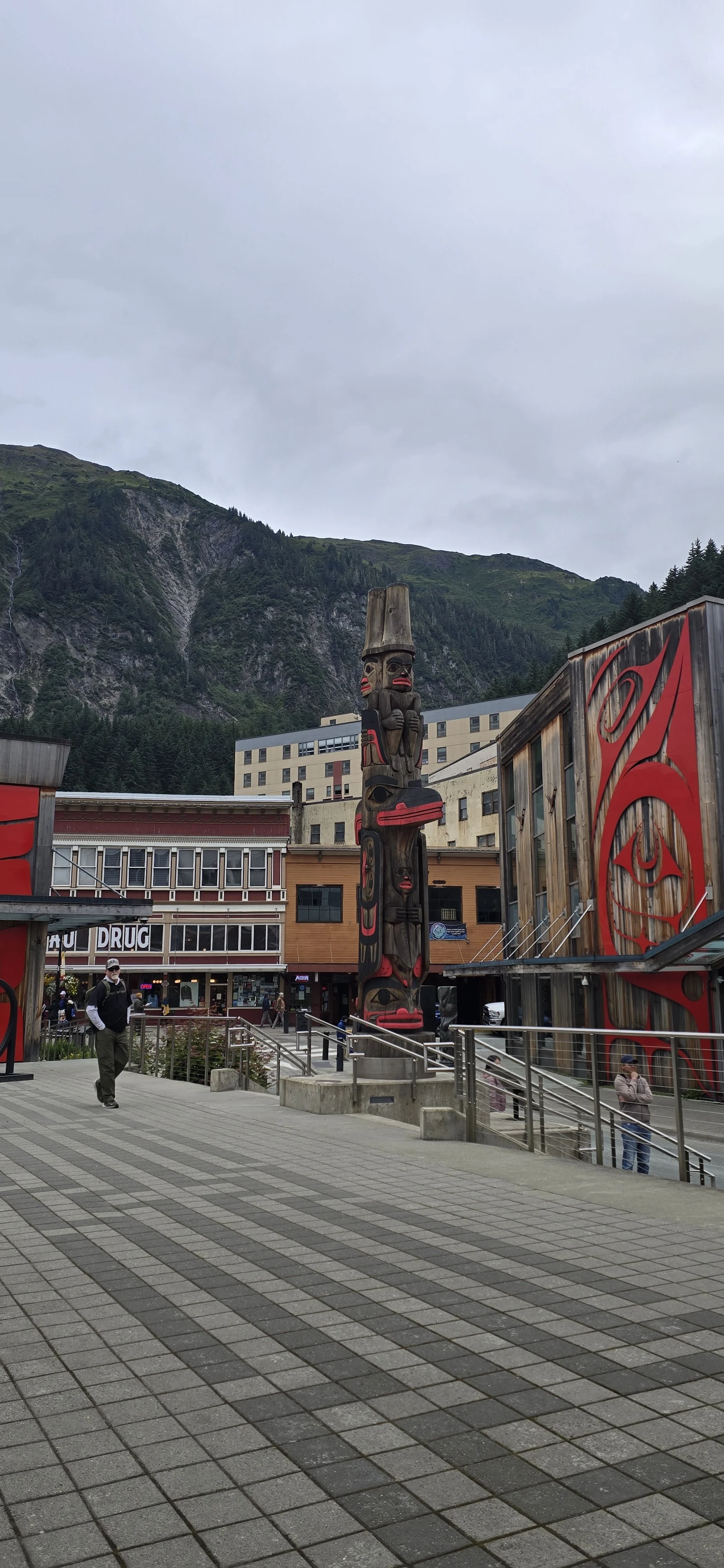 Downtown area with a totem pole sculpture, buildings, and people walking. Mountain backdrop and cloudy sky.