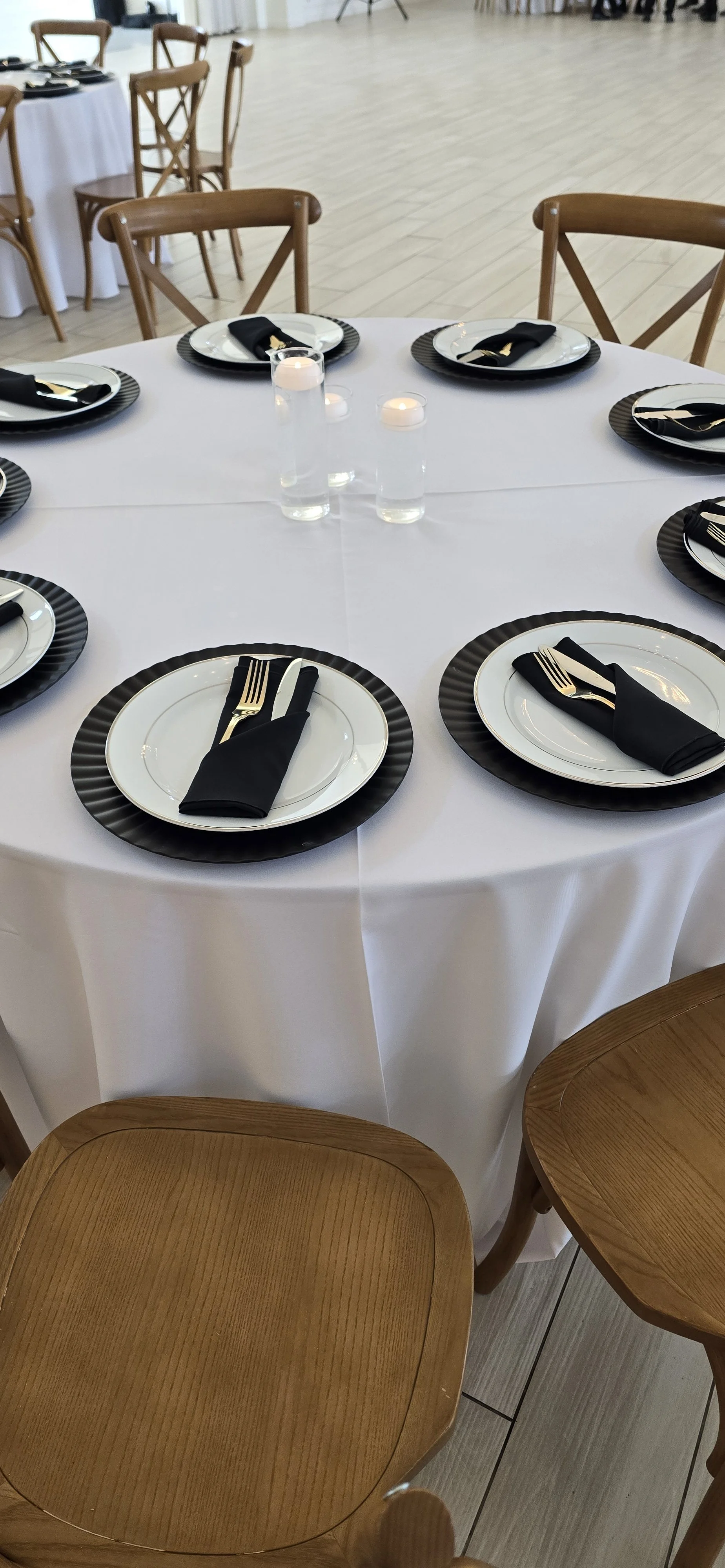 Round dining table set for a formal event with white tablecloth, black and white plates, silverware wrapped in black napkins, and small tealight candles in glass holders. Some chairs are visible around the table.