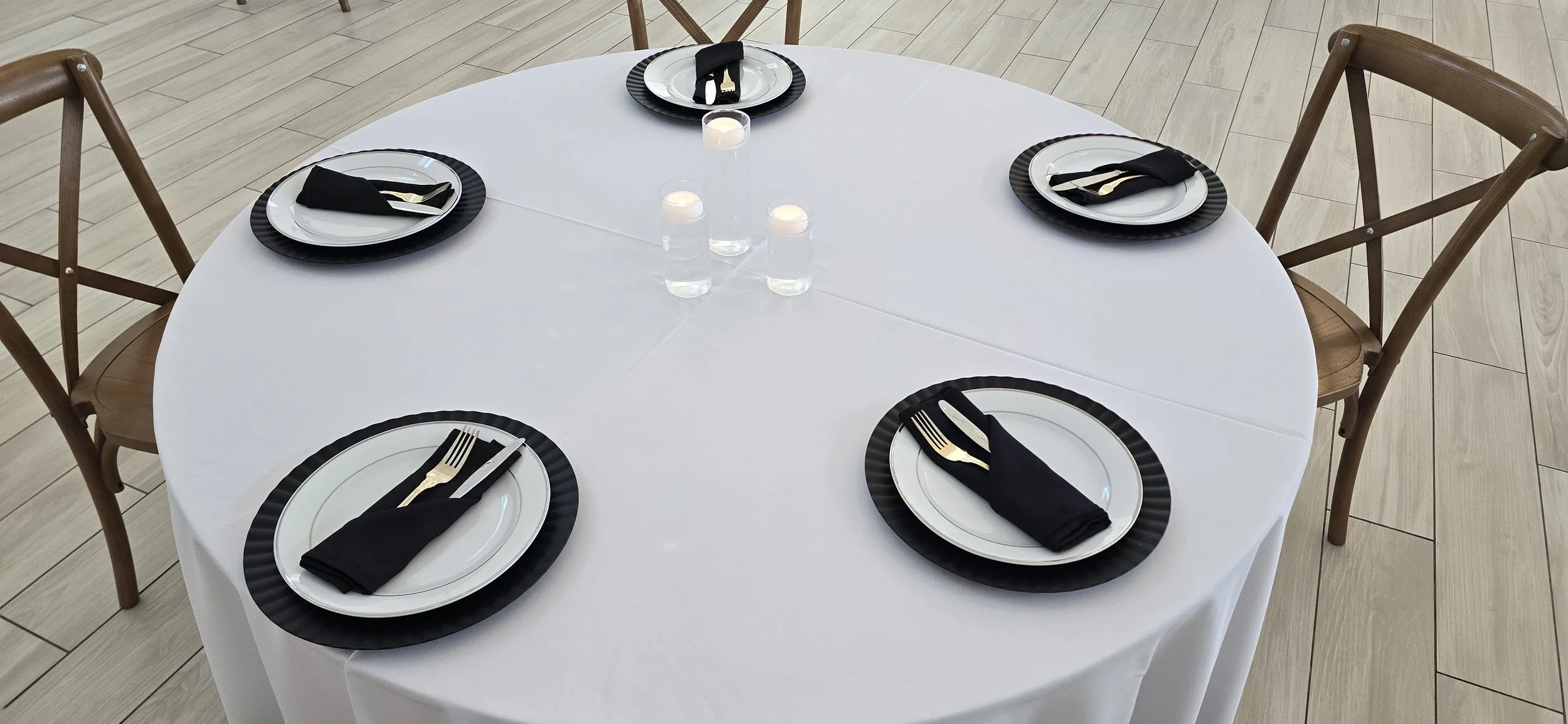 Table set for six with white tablecloth, black and white plates, gold utensils wrapped in black cloth napkins, and three lit candles in the center, chairs around it, on a light wood floor.
