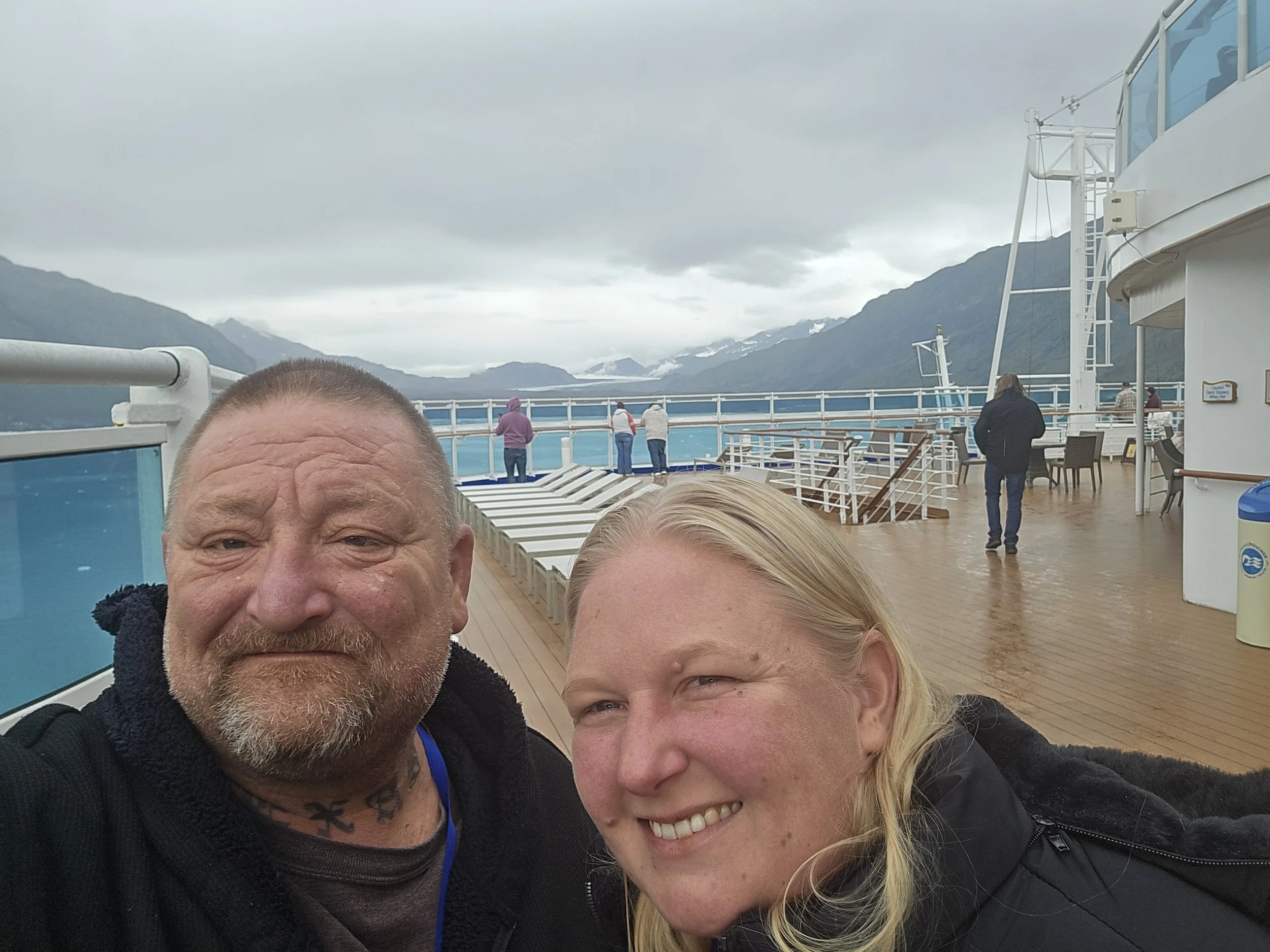 Couple taking a selfie on the deck of a Princess cruise ship in Alaska, with misty mountains and glacier scenery in the background.