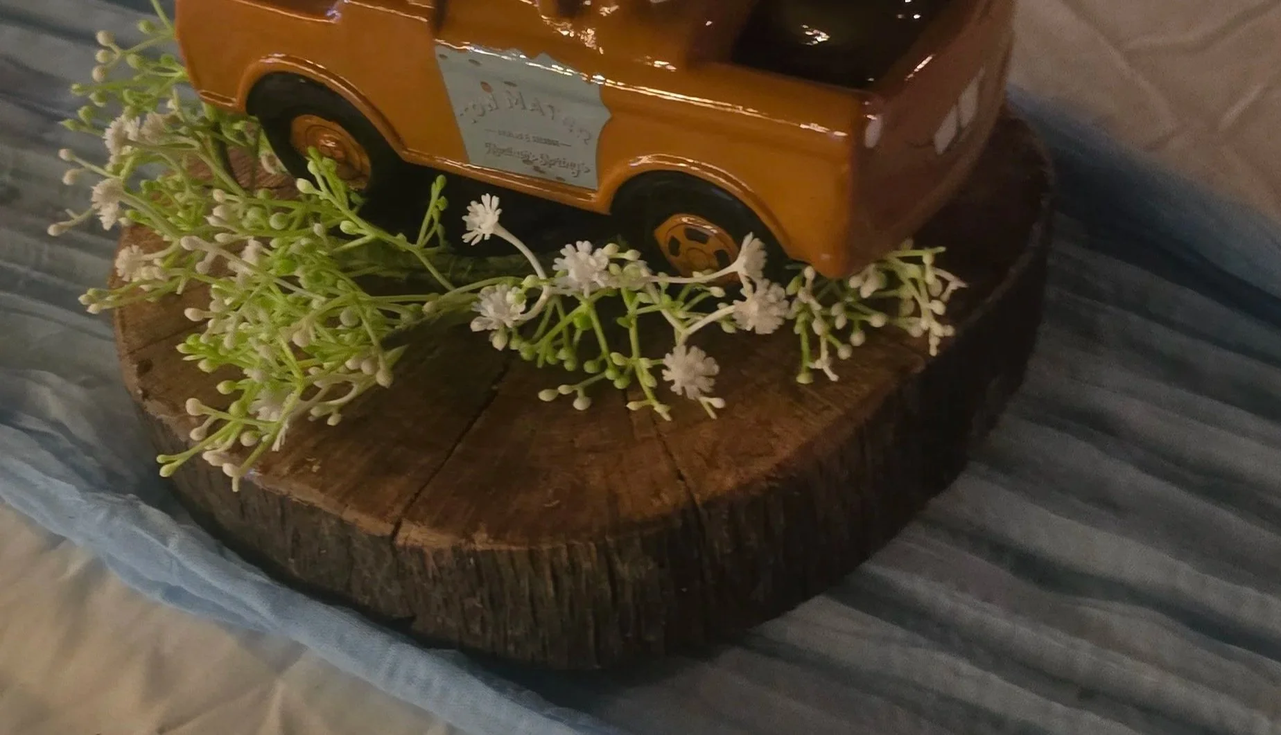 A small wooden table with a round top, decorated with a small model orange vehicle and a cluster of white flowers with green stems. The table is positioned on a blue, textured surface.