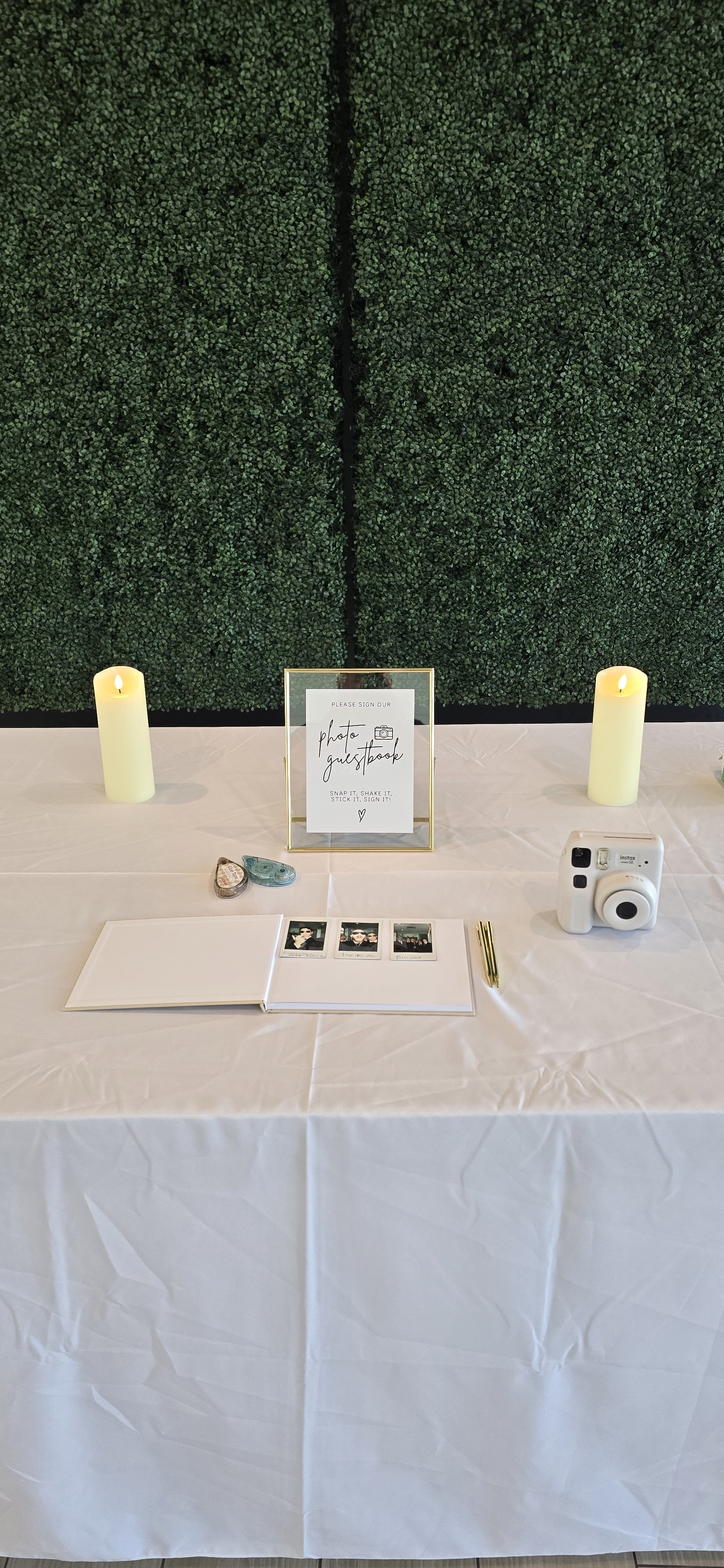 Table setup with a sign, polaroid pictures, a pen, two candles, a guestbook, and a camera against a green hedge background, likely for a guestbook or photo sign-in at an event.