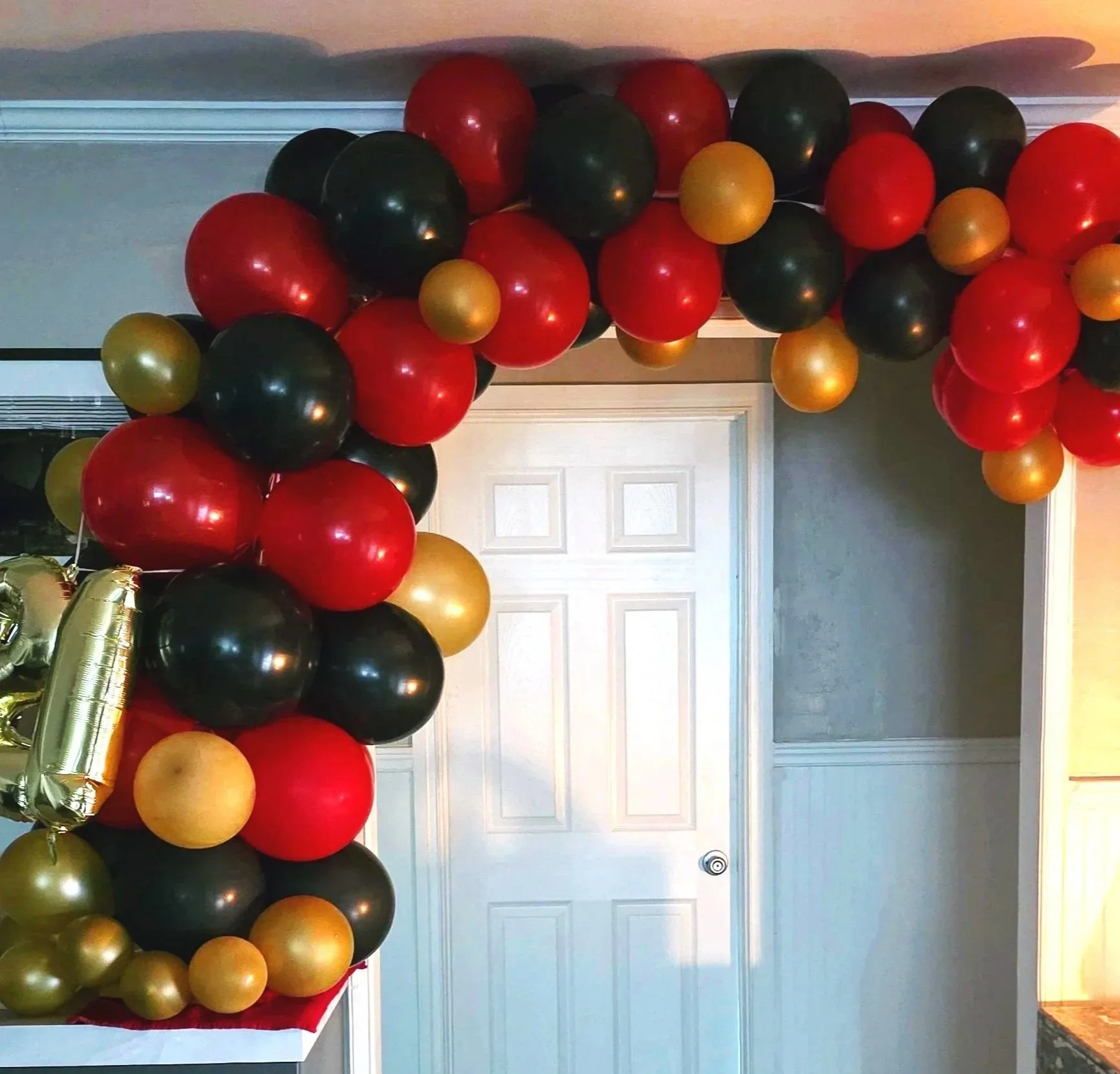 Colorful balloon arch in red, black, and gold decorating a room with a white door and light blue walls.