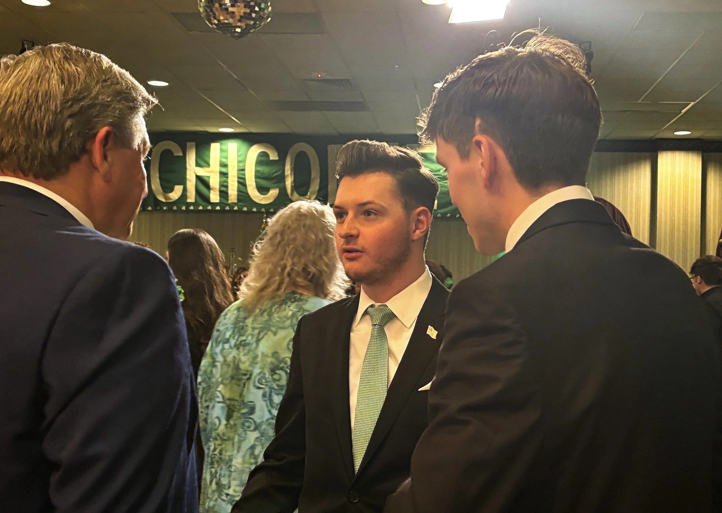 Three men in suits are conversing at an event, with a green and silver banner reading 'CHICOPEE' in the background.