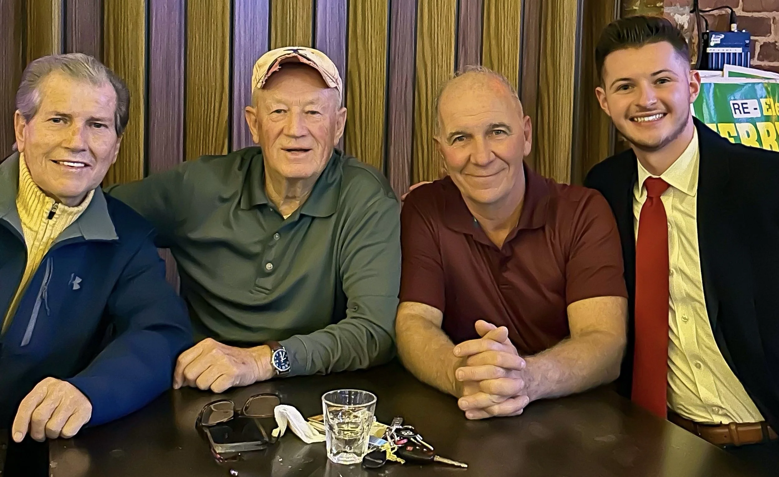 Four men seated together, smiling, in a casual setting, with wood-paneled background, glasses, and keys on the table.