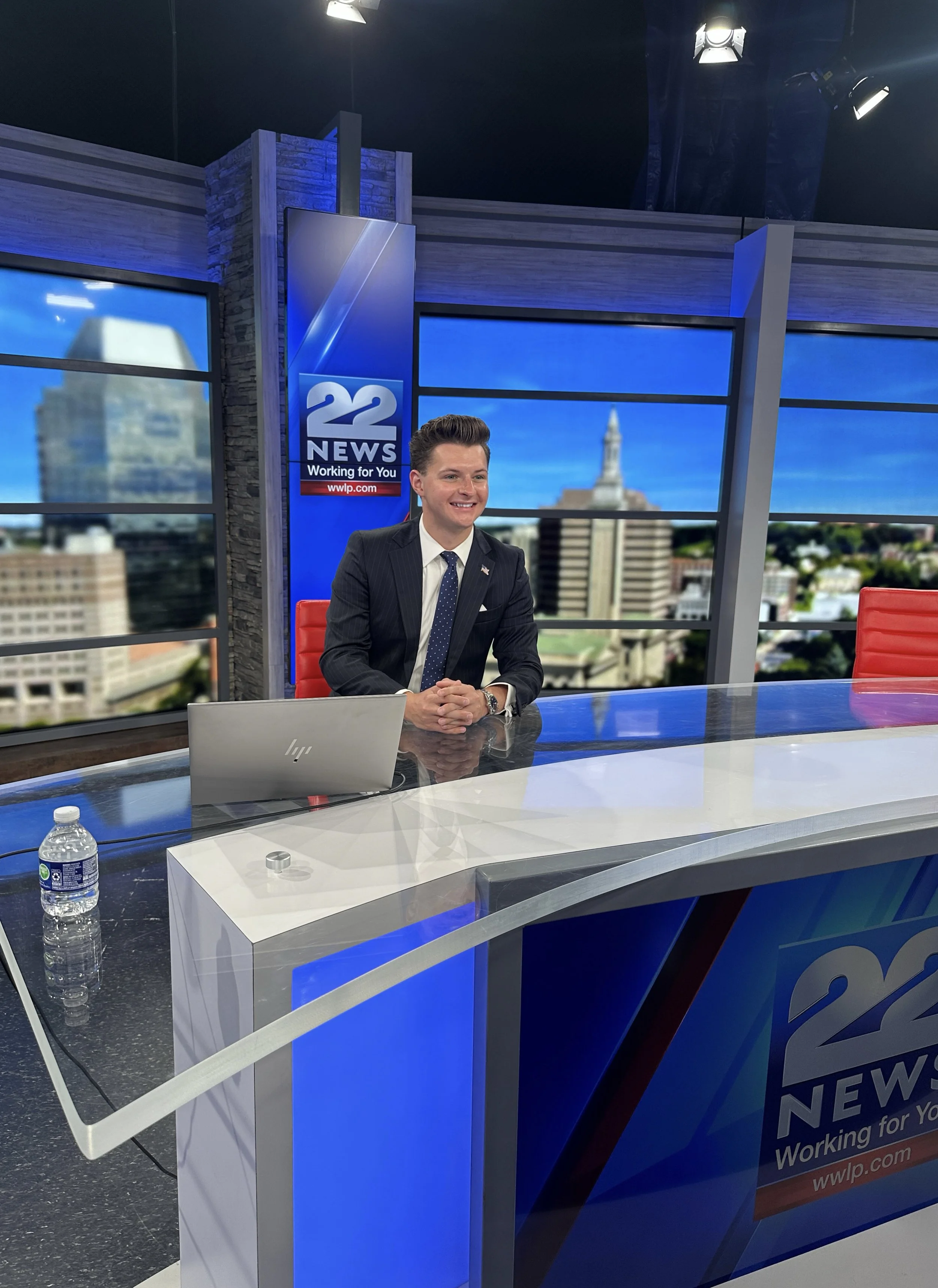 News anchor sitting at a television studio desk with a microphone and laptop in front of him, in a studio labeled '22 News'. Background features large windows with an urban cityscape view.