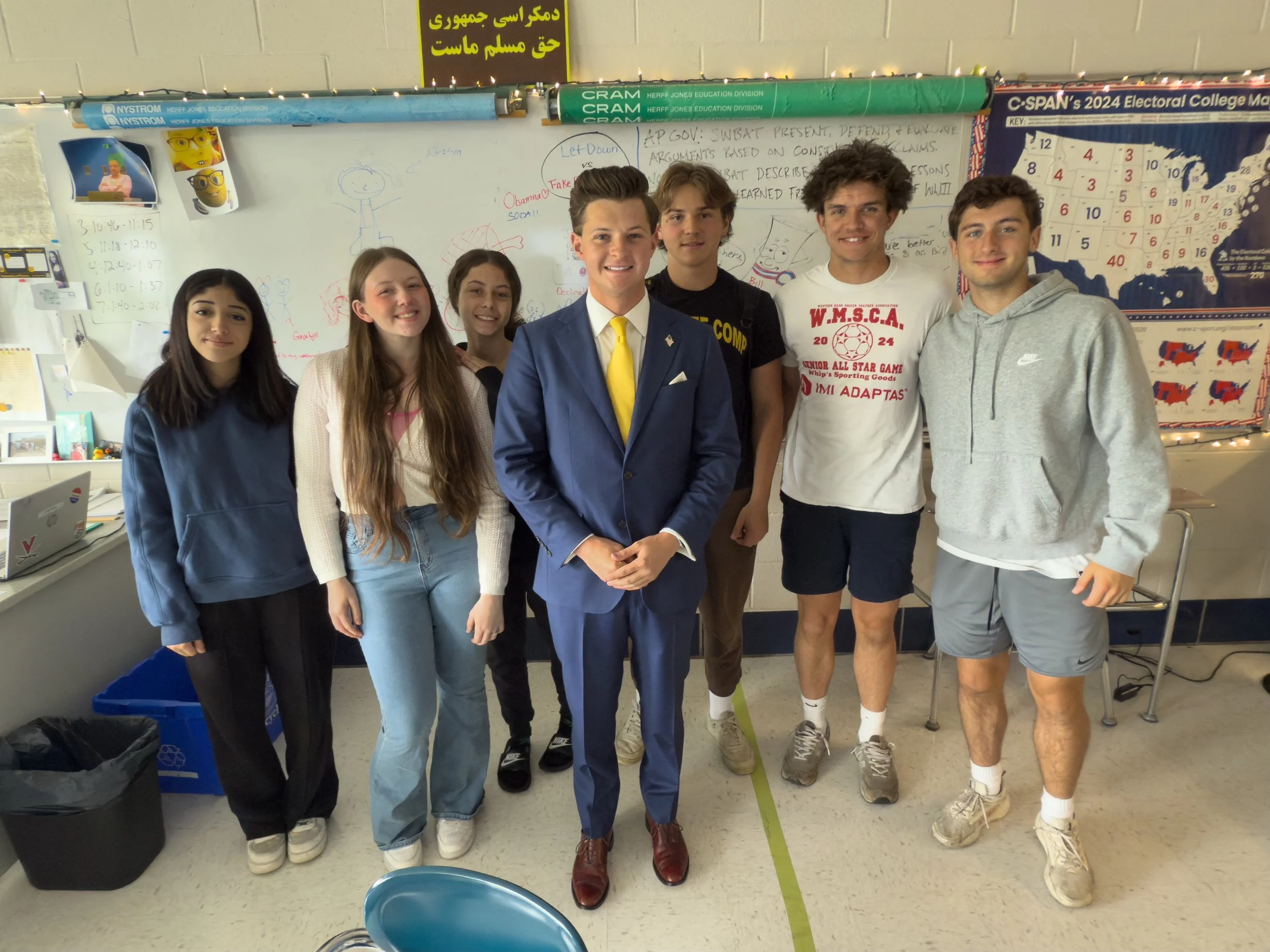 Group of seven students standing in a classroom, some smiling, one in a blue suit with a yellow tie, others casually dressed, with a whiteboard and classroom posters behind them.