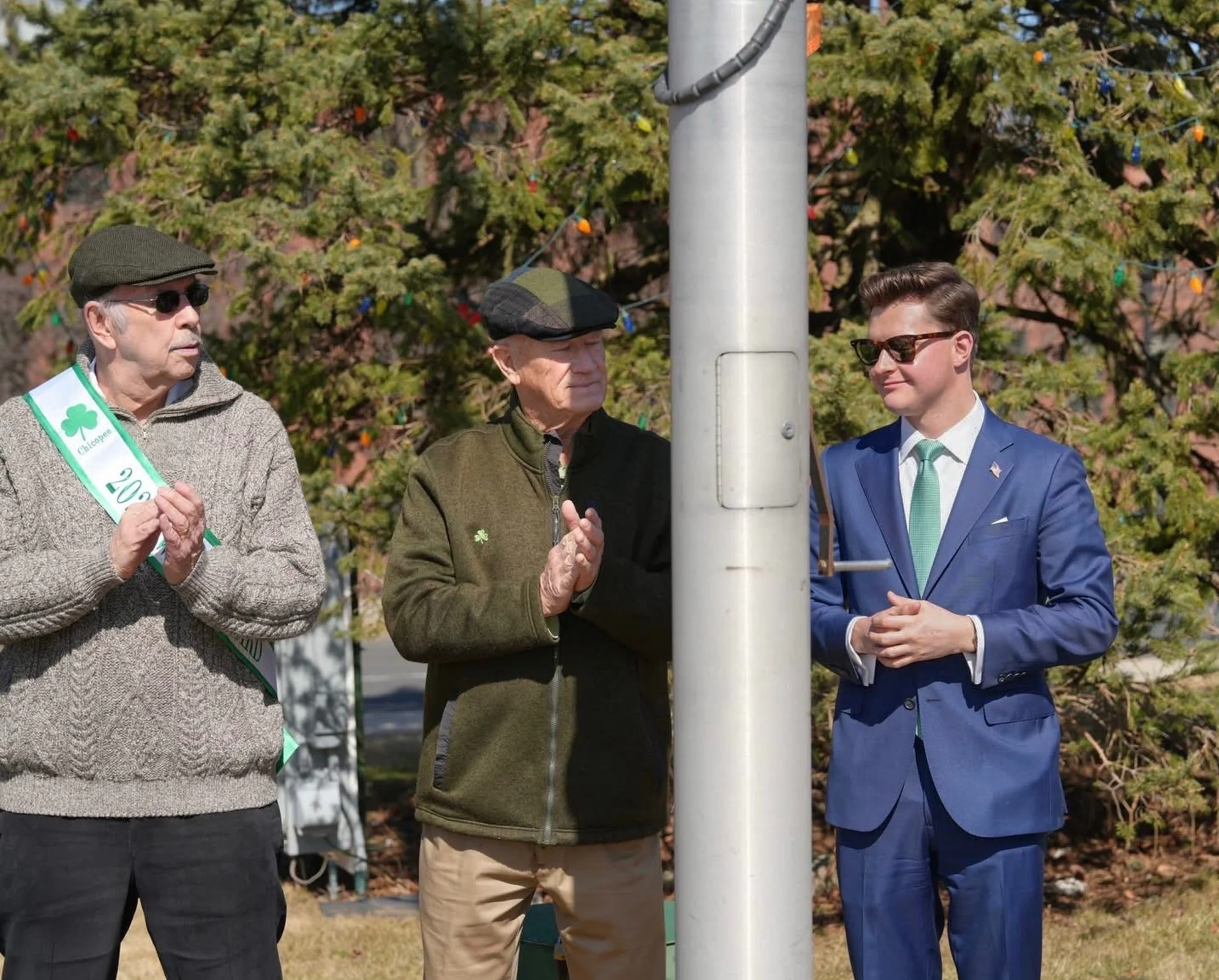 Three men standing outdoors during a St. Patrick's Day celebration, with two of them wearing green and clapping, and the third in a blue suit with sunglasses, standing near a flagpole.