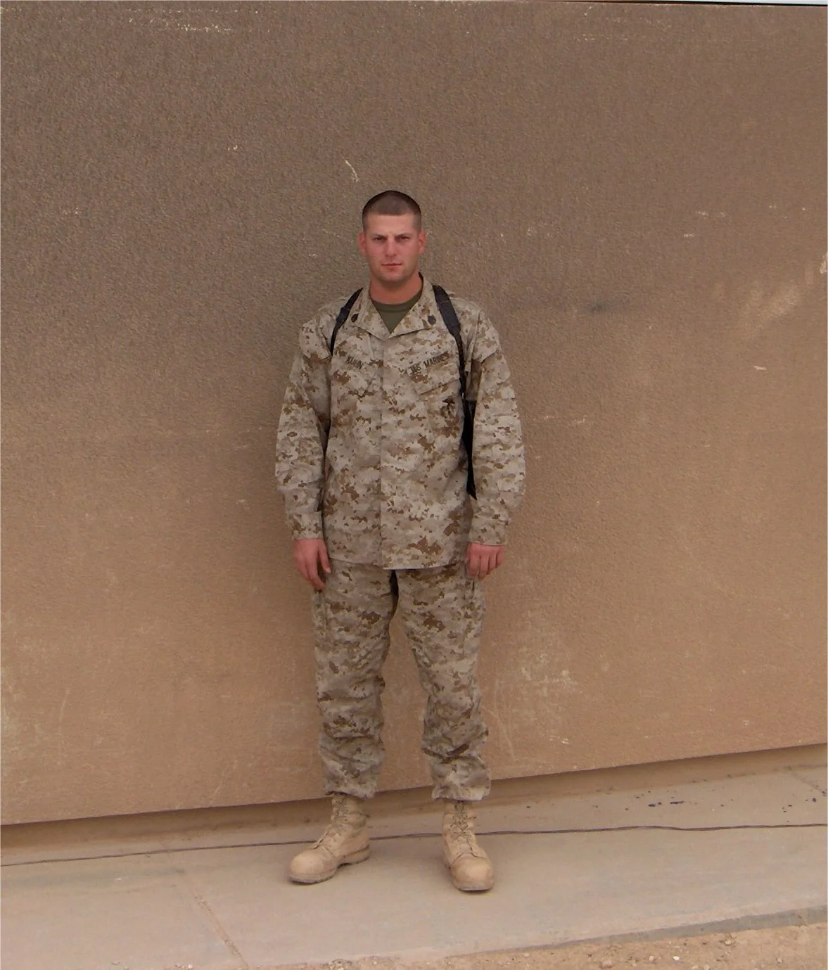 A soldier in camouflage uniform standing against a beige wall, wearing a backpack.