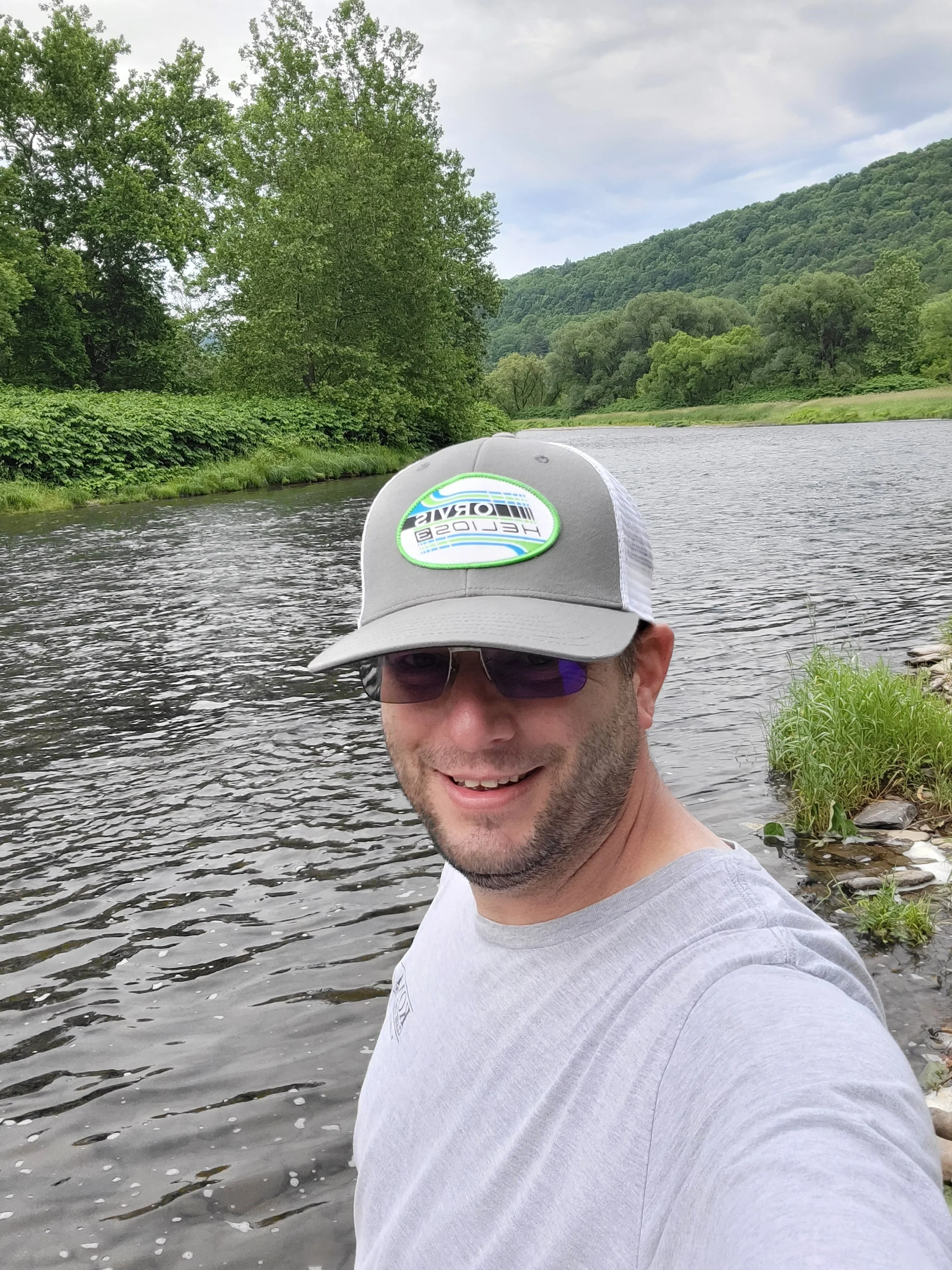 A man with a beard wearing sunglasses, a gray and white cap, and a gray T-shirt taking a selfie by a river, surrounded by greenery and trees in a natural landscape.