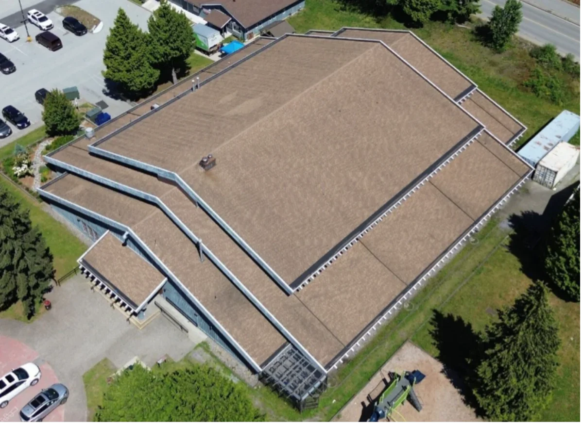 Aerial view of a large building with brown roof, surrounded by parking lot, trees, and green areas.