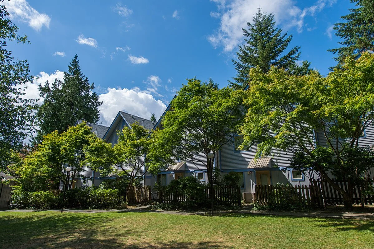 A row of blue townhouses with pointed roofs, surrounded by trees and greenery, under a blue sky with a few clouds.