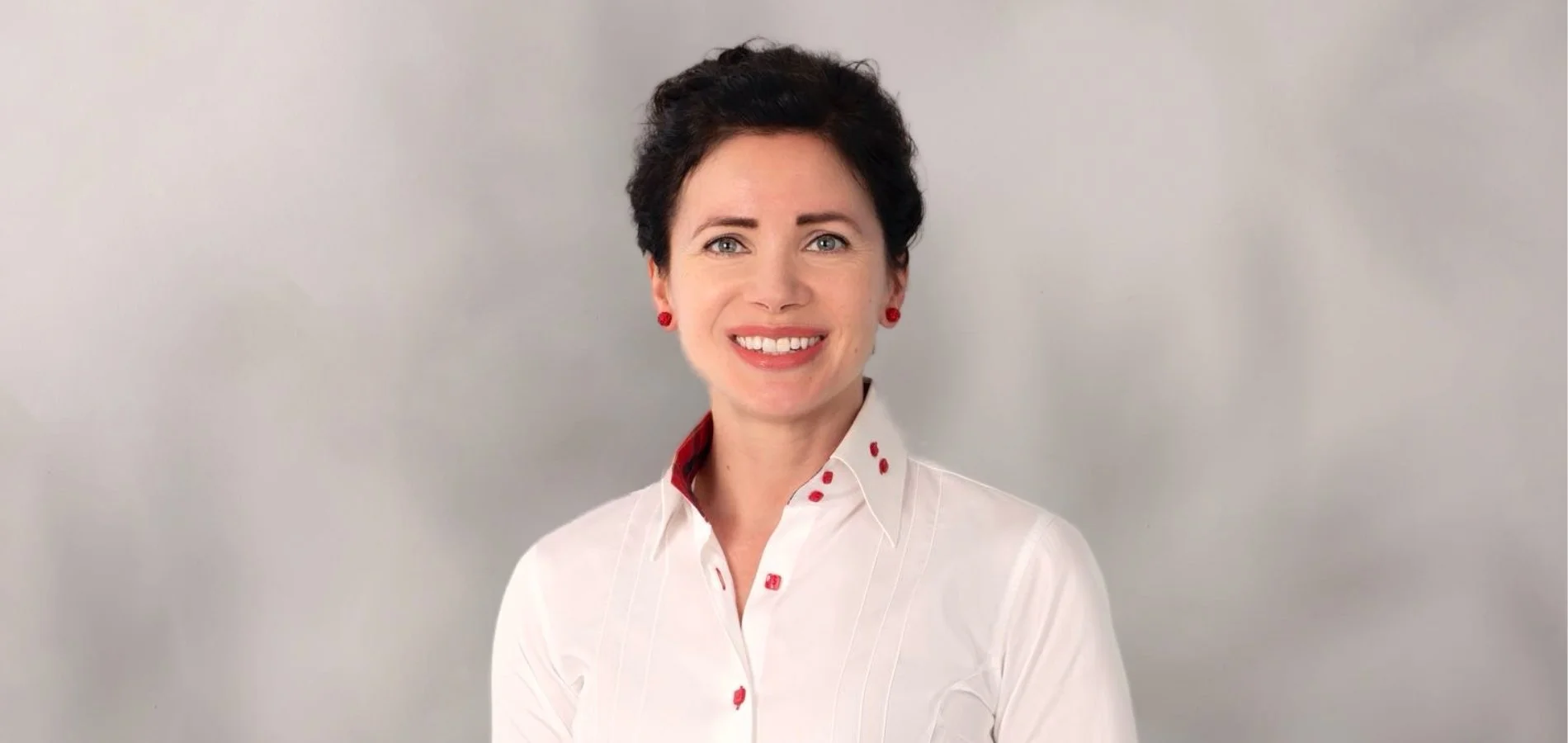 A smiling woman with short dark hair, wearing a white button-up shirt with red accents and red earrings, standing against a light gray background.
