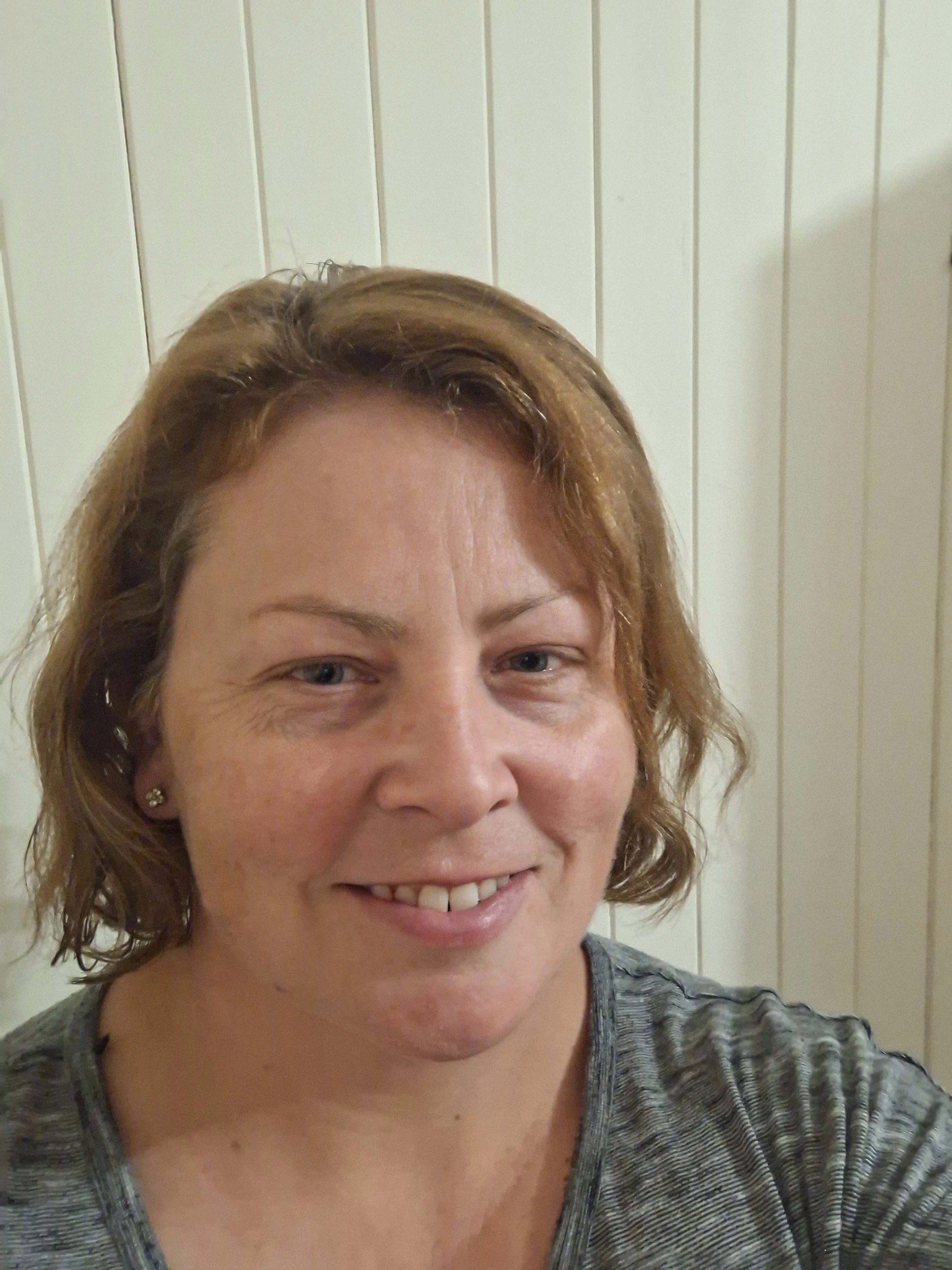 Holly, founder and owner of Riverland Acupuncture, smiling warmly in front of a white panelled wall, wearing a grey top