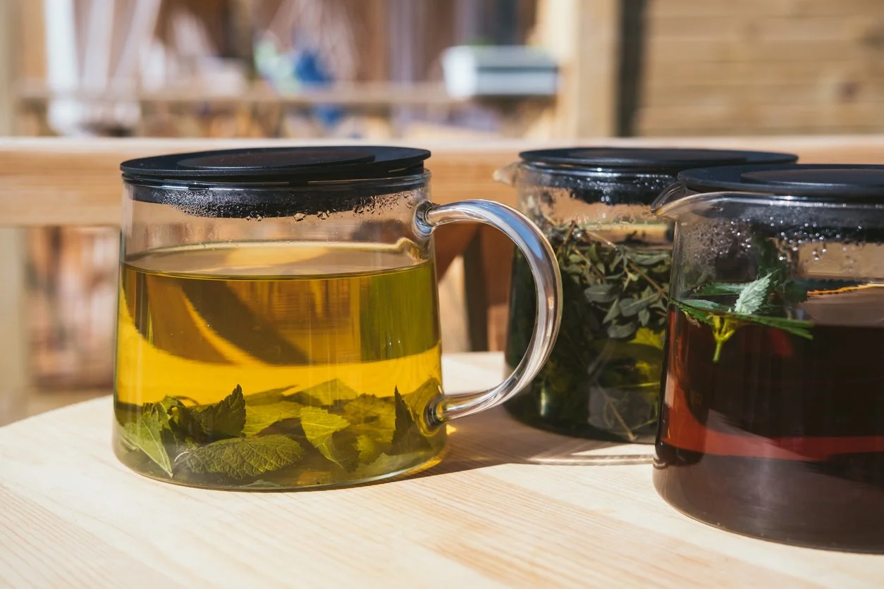 "Three glass teapots filled with different herbal infusions, each containing visible loose herbs steeping in hot water, sitting on a wooden table in natural sunlight