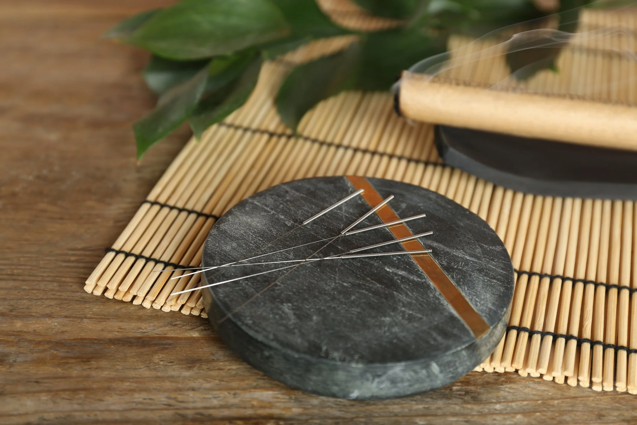 Set of acupuncture needles neatly arranged on a round slate plate with a copper inlay, placed on a bamboo mat over a rustic wooden surface, evoking a sense of tradition and natural healing
