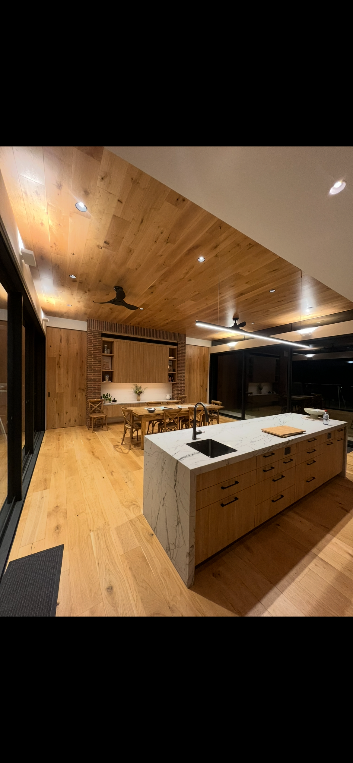 Modern kitchen with wooden floors and ceilings, featuring a marble island with a sink, wooden cabinets, and a dining area with a table and chairs.