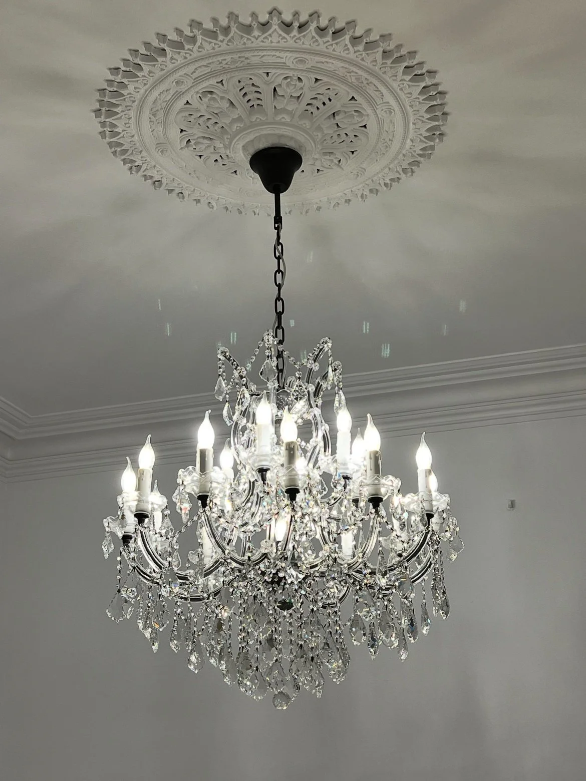 Elegant crystal chandelier with lit candles against a white ceiling featuring an ornate decorative medallion.